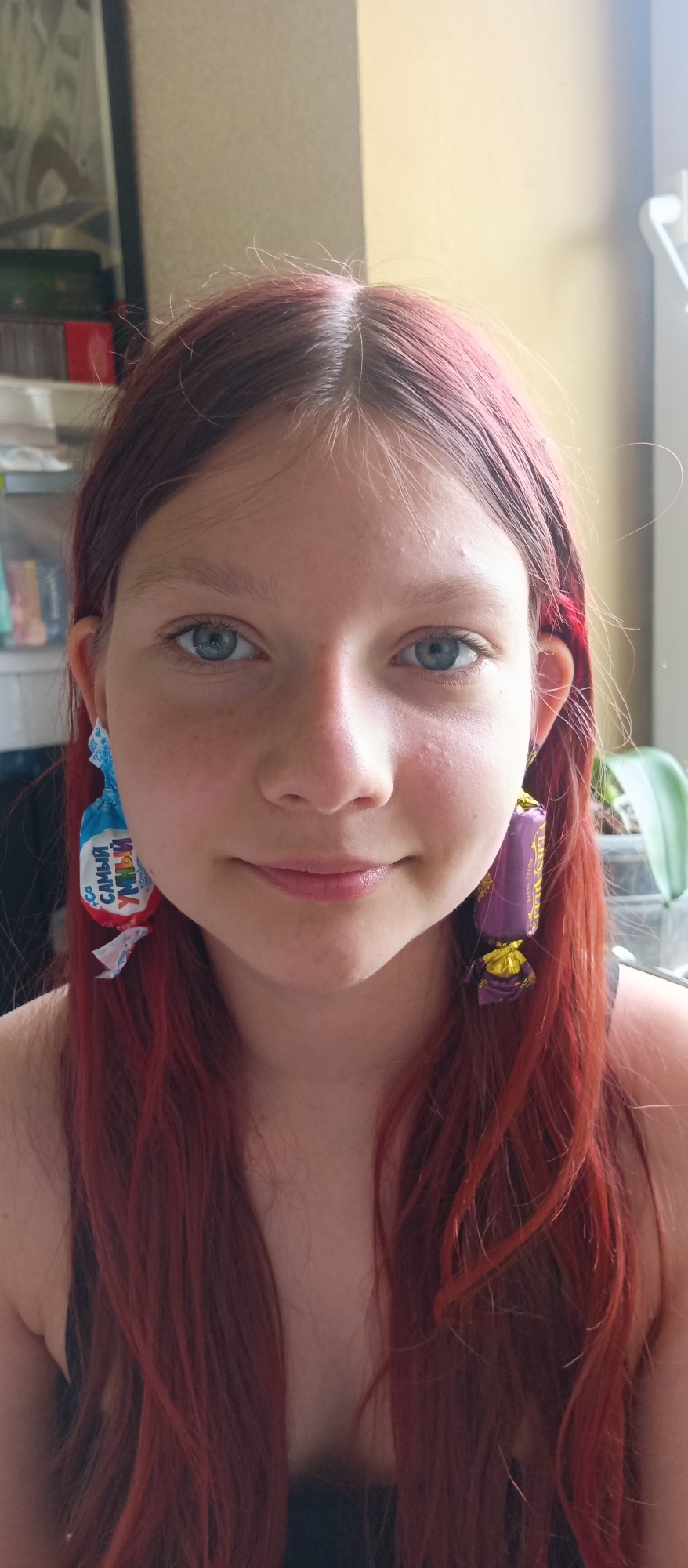 A close-up shot depicts a young woman with reddish-brown hair and blue eyes, centered in the frame. She is wearing earrings crafted from candy wrappers. The left earring is blue and white with Cyrillic text, while the right is purple and gold. She is smiling slightly, and her face is well-lit.
Behind her, the background includes a window to the right with an orchid plant visible. There is a shelf to the left with various items. The city is unspecified, and the setting appears to be indoors.