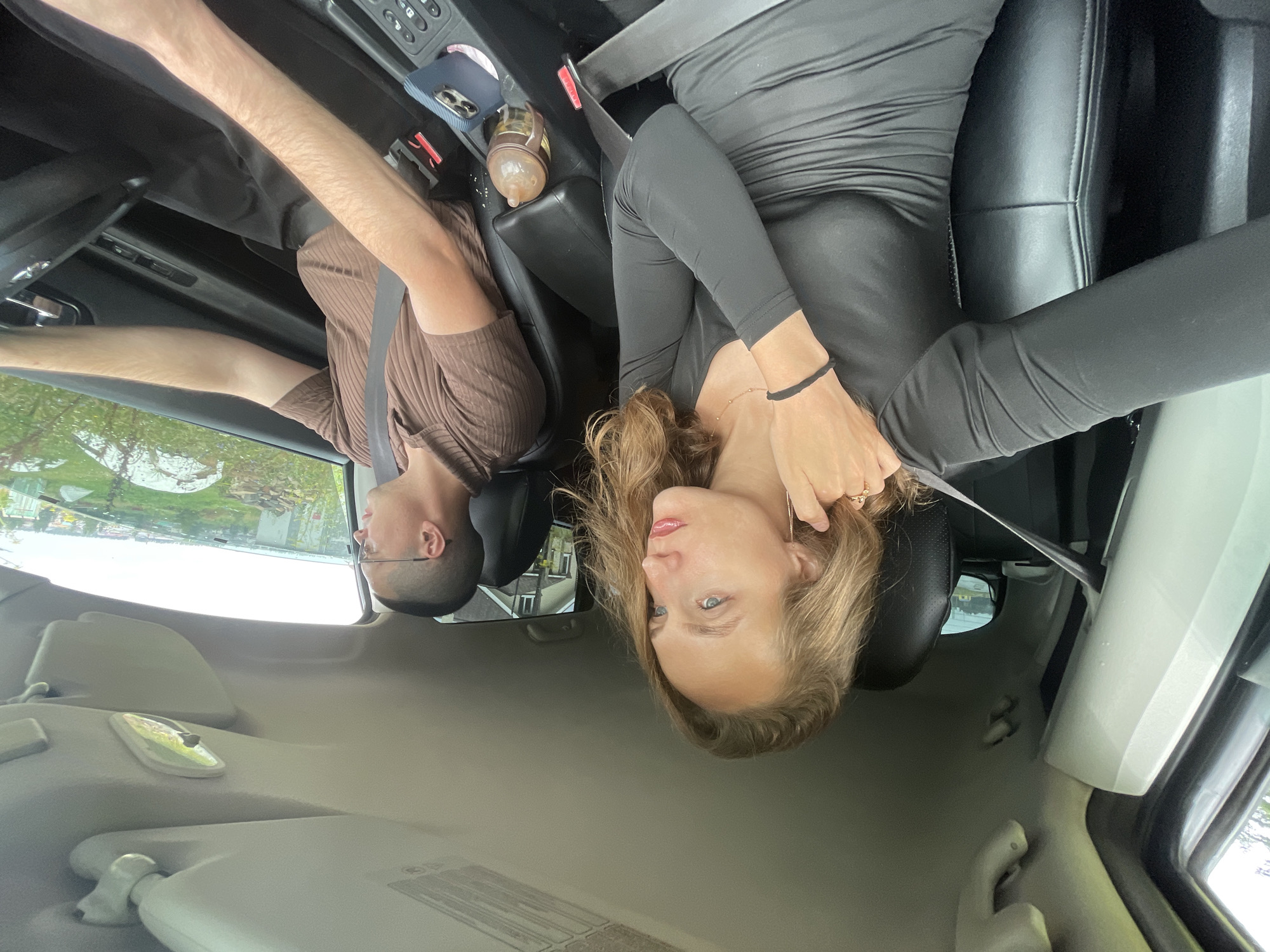 An image shows two individuals inside a vehicle. The woman in the passenger seat is positioned on the left side of the frame, wearing a long-sleeved black dress and looking out the window. She has blonde hair and is touching her face. The man in the driver's seat is on the right side, wearing a brown collared shirt and glasses, and is operating the vehicle. The interior of the car is visible, including the dashboard, seats, and a cup holder containing a baby bottle and a mobile phone. The exterior scene through the windows suggests a rural or suburban environment with greenery and buildings. The image was taken on a cloudy day. This is a candid shot taken from the perspective of someone in the back seat or using a wide-angle lens.