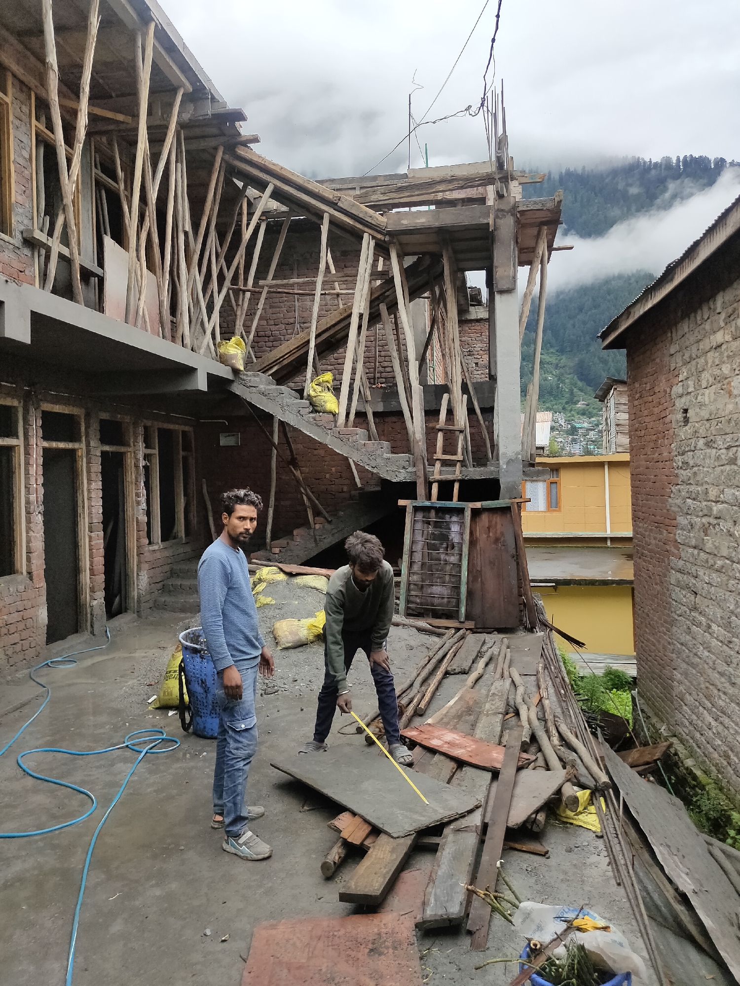 In Siyal, India, an overcast day frames a construction site. Two men are actively engaged in work. One man, wearing a blue sweater and blue jeans, stands near a blue plastic bucket and a coiled blue hose, looking towards the camera. The other man, in a green sweater and dark pants, is bent over a large flat piece of material on a wooden platform, holding a yellow measuring tape.
The scene is characterized by a multi-story building under construction, evident by the exposed brickwork, scaffolding made of wooden poles, and concrete elements. A staircase with concrete steps leads upwards, supported by more wooden scaffolding. Yellow sacks, likely containing building materials, are visible on the stairs and ground. In the background, other buildings and a hillside covered in dense green trees and mist are discernible, suggesting a mountainous or hilly terrain. The ground is a mix of concrete and loose gravel, littered with construction debris including planks of wood and metal rods. A blue plastic bucket containing some plant material and a white plastic bag is in the foreground.