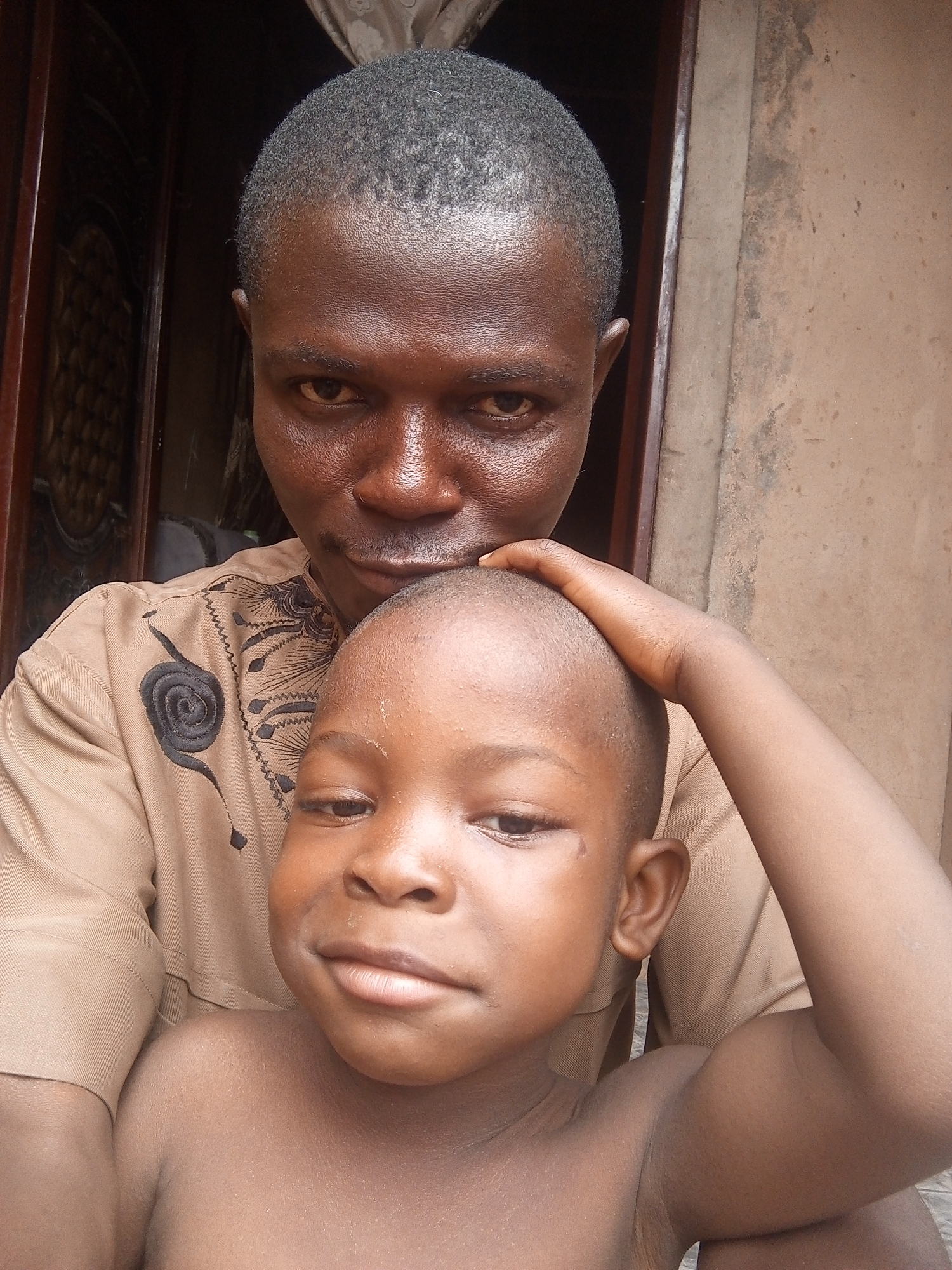 A medium close-up photograph shows an adult man and a young boy, likely a child, in an outdoor setting. The man is standing behind the boy, with his left hand resting on the child's head. The man has dark skin, short cropped hair, and is wearing a light brown collared shirt with a decorative black appliqué on the chest. The boy is shirtless and also has short hair. The wall behind them is a textured, light tan color, suggesting an outdoor setting in a building or compound. A partially visible door is on the left side of the frame. The overall setting appears simple and unadorned, possibly in a residential area. The location is Tempure, Nigeria.