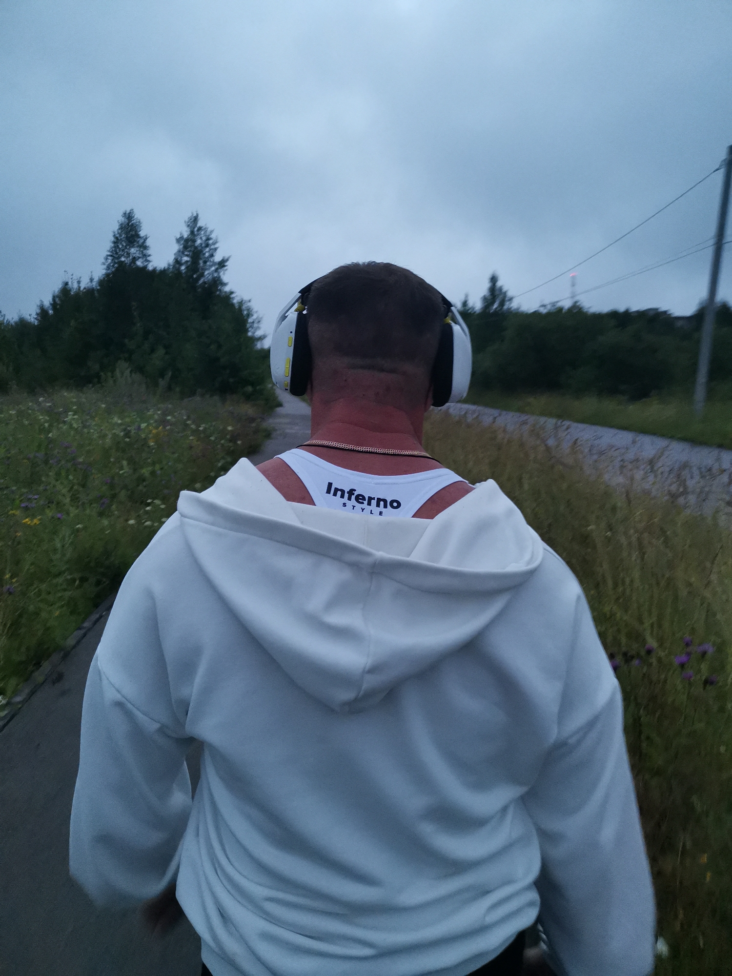 The image shows a person from behind, walking on a paved path. The person is wearing a white hooded sweatshirt and white headphones. A white tank top with black lettering is visible underneath the sweatshirt. The person has short hair and a gold chain around their neck. The environment appears to be outdoors, with tall grass and trees on the side of the path. The sky is overcast and appears to be a cloudy day. In the distance, a tower with a red light can be seen. The overall scene suggests a person walking or jogging in a natural setting, possibly on the outskirts of a city. The location is identified as Kizel, Russia.
