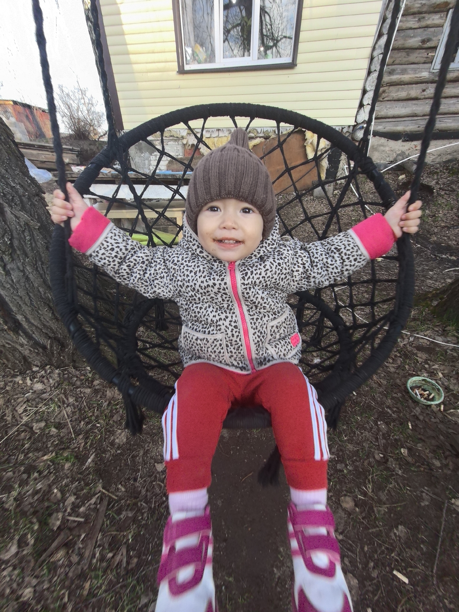 A young child, likely a toddler, is captured in a joyful moment, swinging in a black rope swing outdoors in Noviy, Russia. The child is positioned centrally, looking directly at the camera with a broad, happy smile, indicating delight. A slight smudge is visible around their mouth.
The child is warmly dressed in a brown knitted hat with prominent earflaps, a white and grey leopard-print puffer jacket with a pink zipper and pink cuffs, red track pants with white stripes down the sides, pink socks, and pink and white shoes with velcro closures. Their hands are gripping the black ropes of the swing.
The background shows a residential setting. Behind the child, a light yellow house with horizontal siding and a window reflecting bare tree branches is visible. To the left, the textured trunk of the tree from which the swing hangs is prominent. The ground beneath is covered with sparse brown foliage and dirt, characteristic of late autumn or early spring. Notably, a small green bowl filled with numerous cigarette butts sits on the ground in the lower right. The natural lighting suggests it is daytime with an overcast sky. No discernible text is visible in the image.