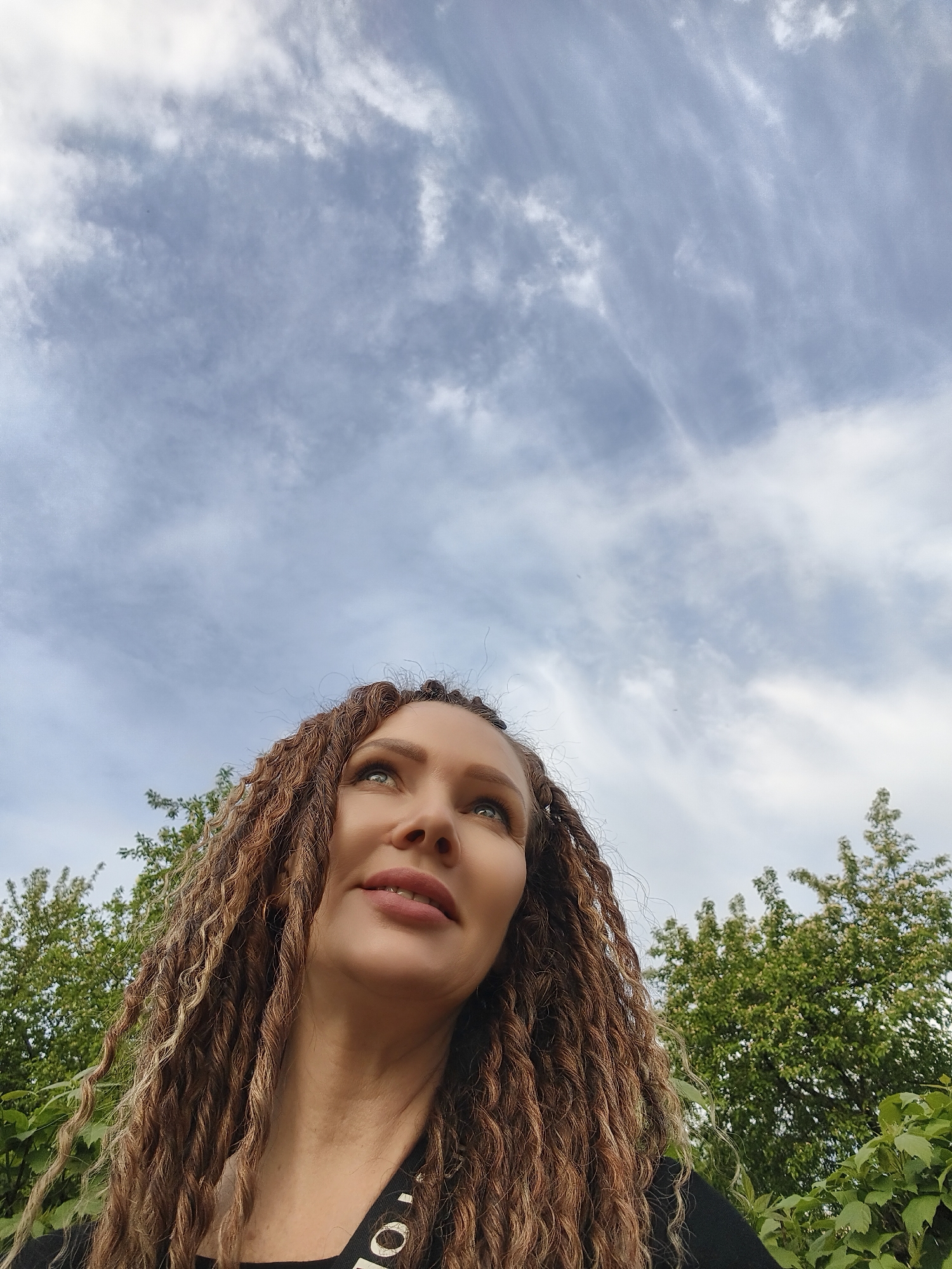 The image is a low-angle shot of a woman with long, brown, dreadlock-style hair looking upwards. She has light skin, green eyes, and is wearing a black top with white writing visible on the strap. The background consists of a cloudy sky and green foliage, possibly trees or bushes. The location is unknown.