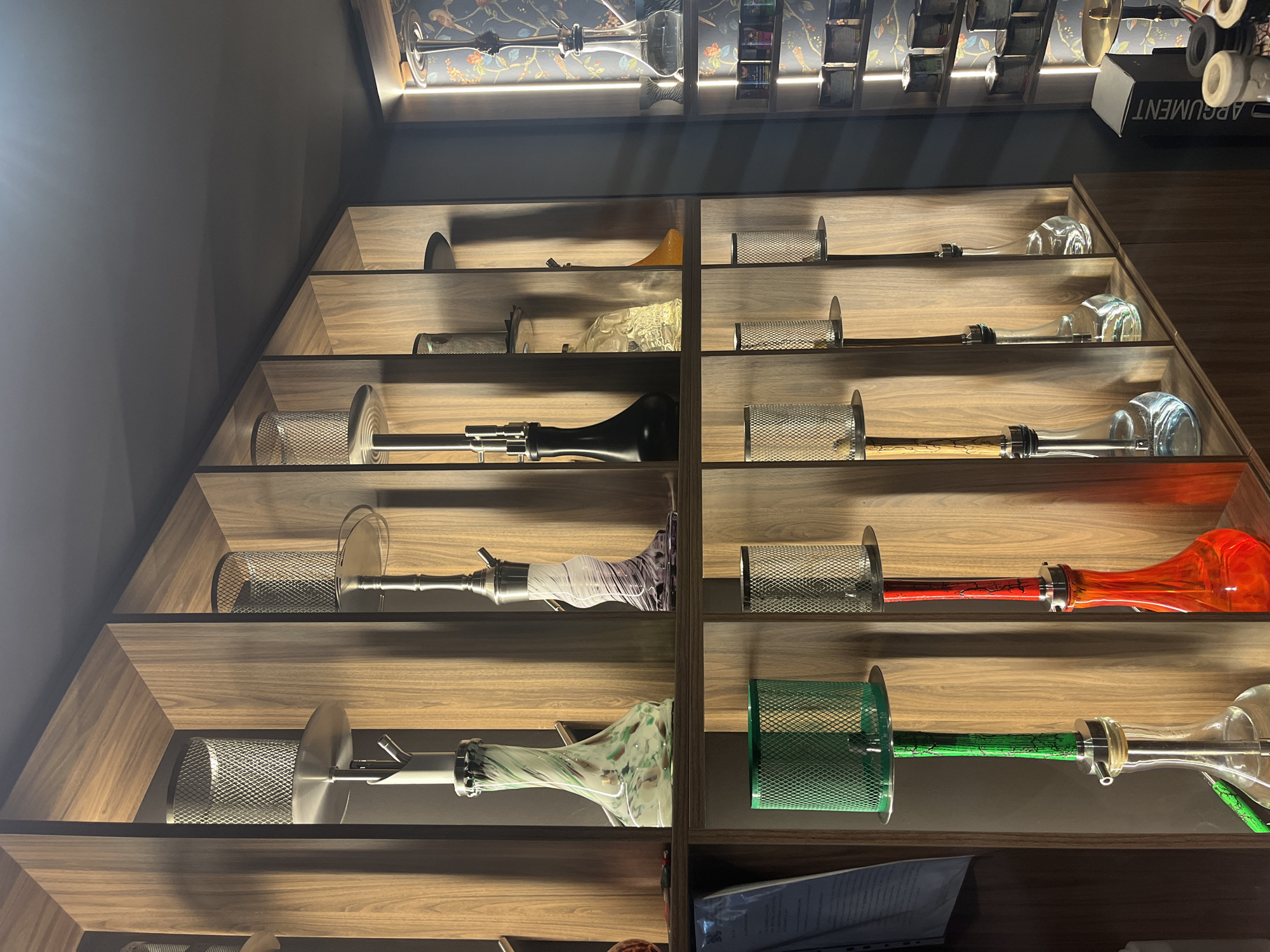 The image shows a retail display of hookahs in what appears to be a shop or lounge. The hookahs are arranged on wooden shelves with a modern, organized design. There are multiple hookahs of various sizes, colors, and styles, with some featuring glass bases and metal stems, while others have colored glass or intricate designs. Some shelves also contain what appear to be accessories or related products. The lighting in the scene is warm and highlights the products. The background shows a patterned wallpaper and more shelves with products. The image does not contain any people or any specific clues to identify the exact location within the city of Plovdiv, Bulgaria, other than what can be inferred from the context of a hookah shop.