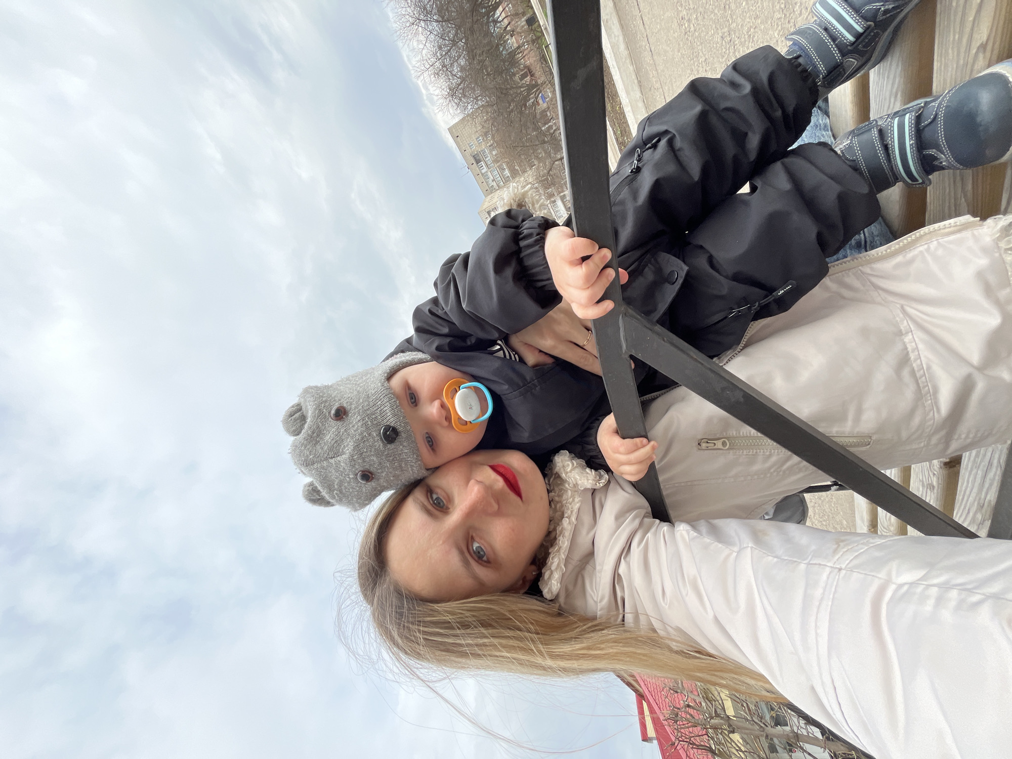 A woman with long blonde hair and red lipstick is holding a toddler wearing a gray beanie and a black winter coat with a pacifier in their mouth. The child is holding onto a black metal railing. The woman is wearing a light beige jacket with a shearling collar. The background includes a cloudy sky, buildings, and bare trees. The location is the city of Chaikovsky, Russia.