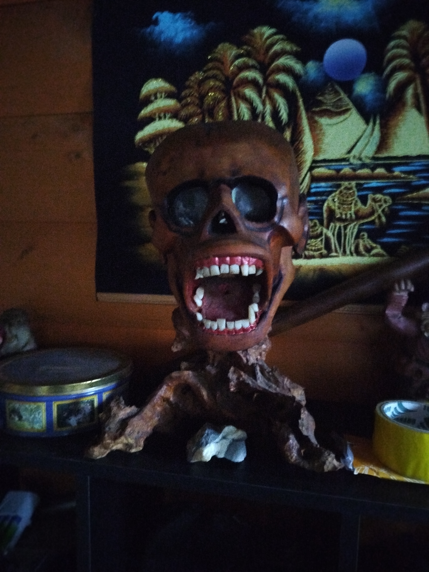 The image shows a carved wooden skull with an open mouth and visible teeth, perched on a gnarled wooden base. The skull sits on a dark shelf or surface alongside a round metal tin and a roll of yellow tape. Behind the skull hangs a dark tapestry depicting a desert scene with pyramids, palm trees, camels, and a blue moon. The image appears to be taken indoors. There is no evidence to support the location being in the city of Боровково, Russia, or any information about people, subjects, objects, actions, interactions related to that location.