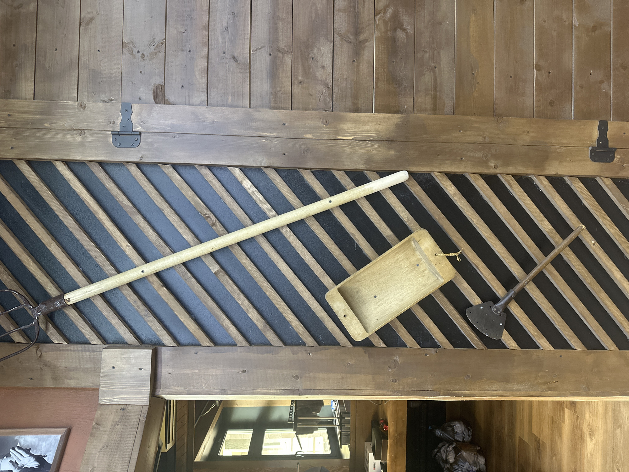 The image depicts a section of a rustic interior wall, likely indoors in Selyatino, Russia. The upper portion consists of vertically oriented wooden planks of varying widths, stained in a medium brown tone. Two black metal hinges are attached to the horizontal plank bordering the top of a panel below.
The panel below features a pattern of alternating diagonal wooden slats in natural and dark grey tones. Several tools are displayed on this panel. A long-handled pitchfork lies diagonally across the slats. To the right, a wooden tray with a small handle hangs by a string. Further to the right is a short-handled spade or shovel.
Below the panel is a wooden beam. The image provides a glimpse into the room below. This room appears to be a storage or utility area with a window and various objects.