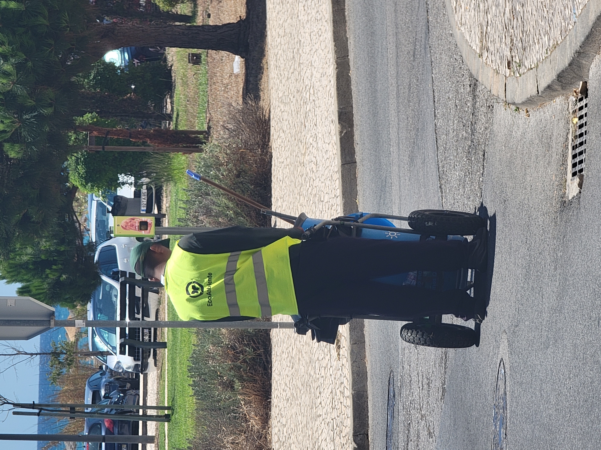 A person wearing a neon yellow safety vest with reflective stripes and the logo "EcoAmbiente" is standing in the street, facing away from the camera. They are also wearing dark pants and a dark cap. The person is pushing a cleaning cart that has two large wheels. The cart appears to be blue and contains cleaning supplies, including a broom with a blue handle. The scene is located in Quarteira, Portugal, likely near the waterfront given the presence of parked cars and distant view of the ocean and beach umbrellas. The street is paved, and a patterned tiled sidewalk runs alongside it. There are trees and vegetation in the background, and a metal grate is visible on the road surface. The lighting suggests it is daytime, with sunlight casting shadows.