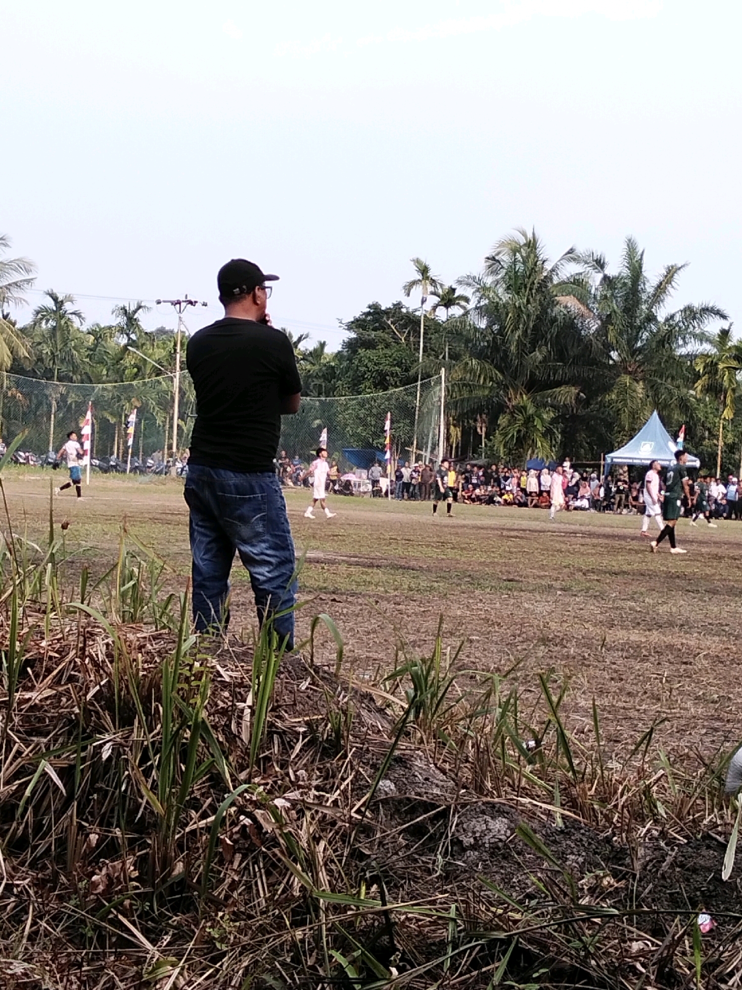 In Pambang Pesisir, Indonesia, a group of people are gathered on a dirt field, observing a soccer game. A man in a black t-shirt and jeans stands in the foreground, facing away from the camera. Several players in soccer uniforms are visible on the field, along with spectators sitting on the grass and standing behind a fence. Palm trees and other vegetation surround the field, and a tent is set up on the right side of the image. The sky is clear and overcast.