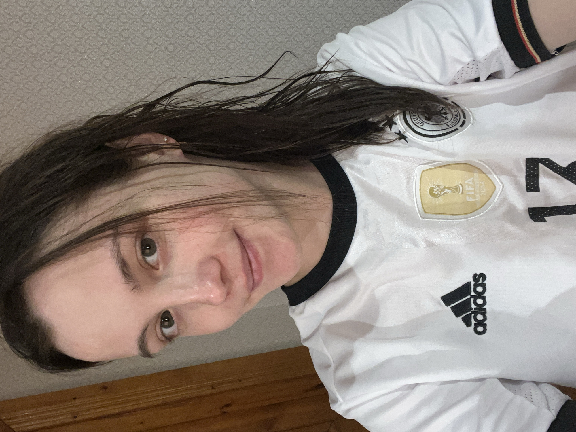 A young adult female with fair skin, dark brown wet hair, and dark eyes is depicted in a close-up, chest-up shot indoors. She wears a white short-sleeved German national football team jersey with a black collar. The jersey features a black Adidas logo, a gold FIFA World Cup 2014 champions badge, and a circular German Football Association crest with three black stars above it. The number "17" is partially visible in black on the lower part of the jersey. Her right ear has a small stud earring. She is looking directly at the camera with a subtle smile. To the left, a vertical wooden door frame or panel is visible. The background to the right consists of a light-colored wall with a repeating, subtle geometric pattern.