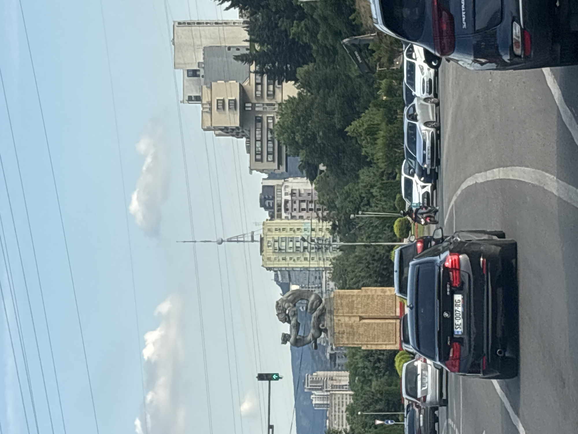 The image depicts a street scene in Tbilisi, Georgia, likely during the daytime given the bright lighting and clear sky with some clouds. The perspective is from a vehicle looking forward, showing a line of parked cars on the right and buildings and trees on the left.
Prominent subjects include several black and white cars, some appearing to be SUVs. A black BMW with the license plate "SE-007-RG" is clearly visible. In the background, there are various modern buildings with multiple windows and balconies, interspersed with lush green trees. A notable landmark is a statue of two figures on a pedestal, situated between the street and the buildings. A tall, slender tower or antenna is also visible in the distance, above the buildings. Power lines crisscross the sky, a common sight in urban environments. A traffic light with a green arrow is also visible.
The scene suggests a typical urban setting with traffic and pedestrian infrastructure. The presence of cars, buildings, and a statue indicates a city environment. The overall mood appears calm and uneventful, likely a regular day in the city. No people are clearly discernible as subjects in the foreground, though they may be present inside the buildings or vehicles. The weather seems pleasant, with a clear blue sky and scattered white clouds. The time of day appears to be late morning or early afternoon due to the bright sunlight.