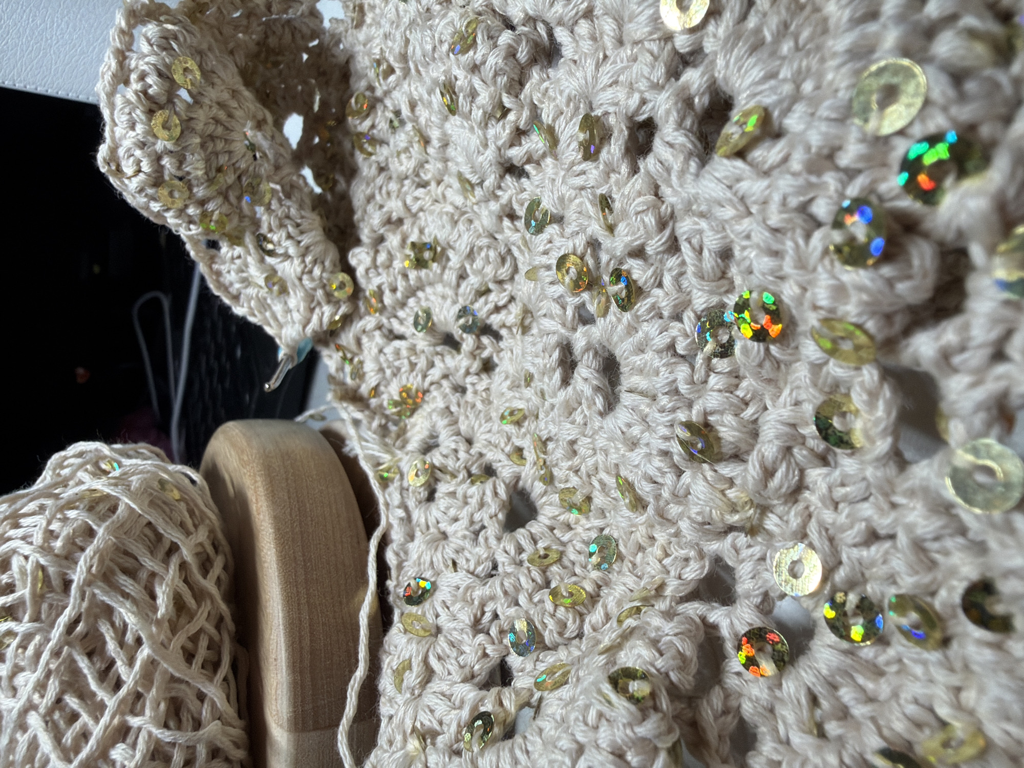 A close-up shot shows a crocheted cream-colored garment, possibly a shawl or top, adorned with gold and iridescent sequins. The crochet pattern has a loose, open weave. A ball of yarn of the same color, also featuring sequins, sits next to a light-colored wooden object, possibly a spool or part of a loom. The background is partially obscured and dark. There is no information to suggest the location of Chaikovsky, Russia.