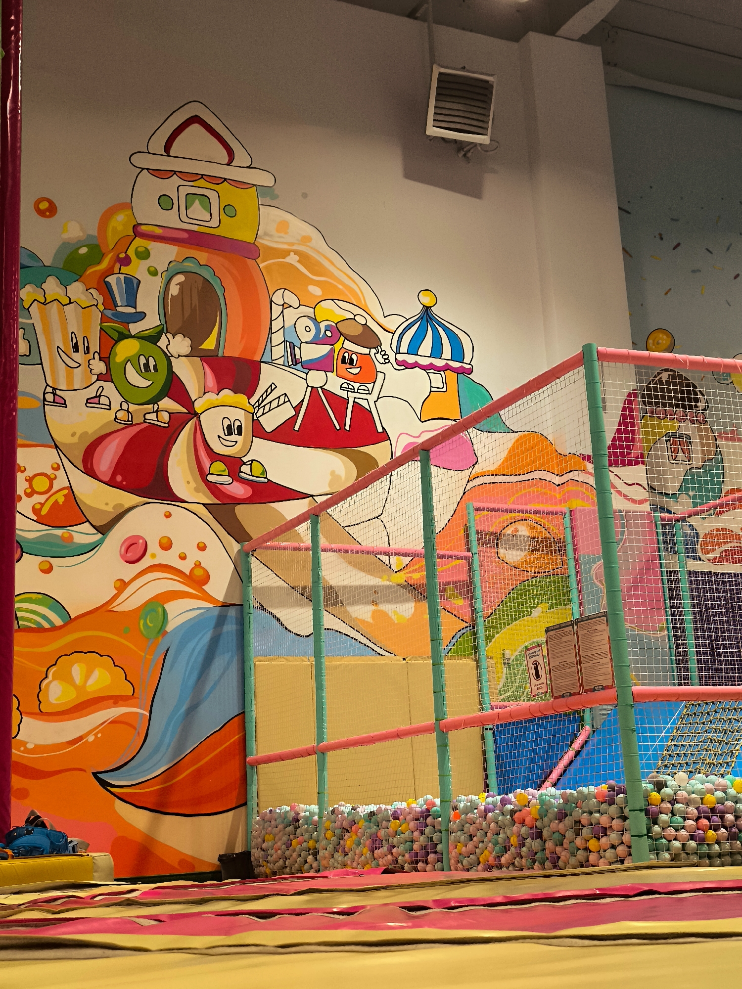The image depicts the interior of a brightly colored indoor playground, likely a children's entertainment center. The walls are adorned with a vibrant mural featuring cartoon characters and whimsical, candy-themed elements, including a house, popcorn, an apple, and other cheerful figures. In the foreground, a padded play area with yellow and pink mats is visible, leading towards a netted enclosure. This enclosure contains a ball pit filled with numerous multi-colored plastic balls. The play area is equipped with protective netting and supports, colored turquoise and pink, indicating a safe environment for children. A heating or ventilation unit is mounted on the upper wall. The overall impression is one of a fun and engaging space designed for recreation. The provided crops confirm the details of the mural and the play area.