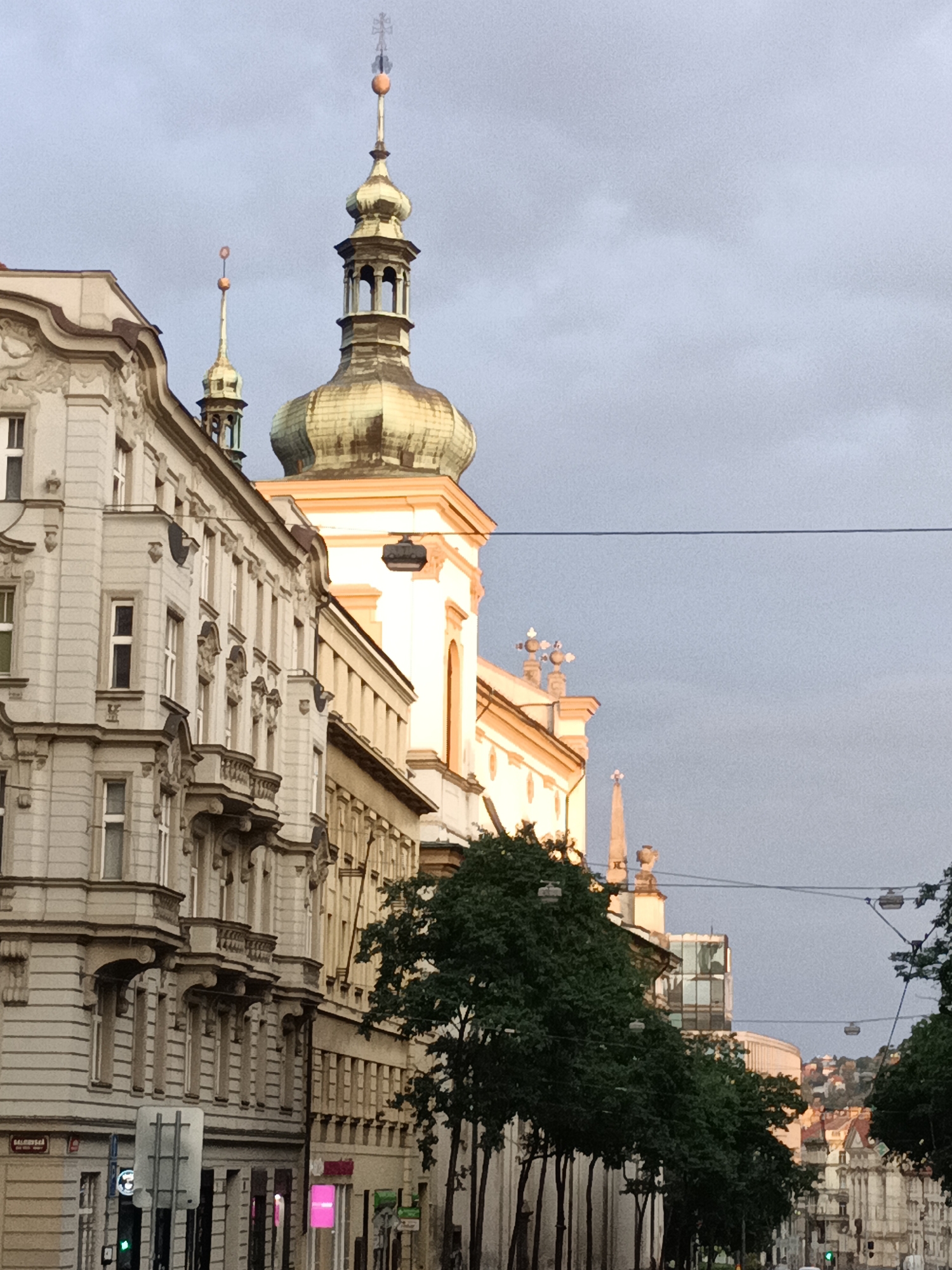 The image depicts a street scene in Prague, Czechia, captured on a cloudy day. The foreground is dominated by the facades of several historic buildings, characterized by ornate architectural details, multiple windows, and balconies. A prominent golden-domed church tower with intricate detailing and a cross at its apex rises above the surrounding structures. Adjacent to it, another smaller, similar tower is visible. Trees line the street, their green foliage contrasting with the muted tones of the buildings and the sky. Power lines are strung across the upper portion of the image. In the background, more buildings and distant hills can be seen. The street appears to be a commercial area, with signs and shop fronts visible, including a bright pink sign. There are no discernible individuals in the frame.