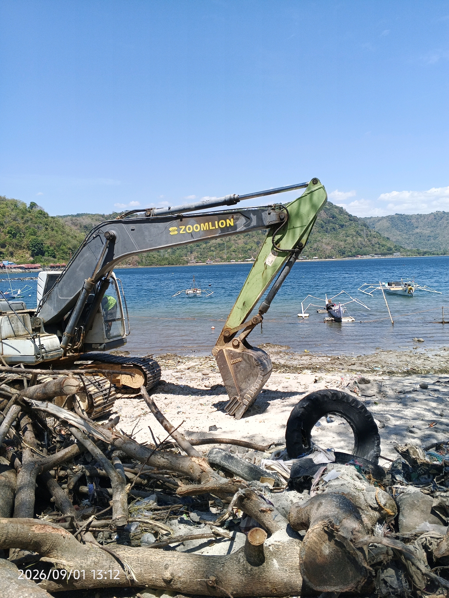 An excavator, with the brand name ZOOMILION visible on its arm, is situated on a sandy beach in Kupa, Indonesia. The excavator is dark gray and green, with a large bucket attachment. In front of the excavator, a pile of logs and debris, including a tire, is visible. The beach is also littered with trash. The sea is a bright blue and extends to the horizon, where lush green hills form the backdrop. Several small boats are anchored in the water. The sky is clear and blue. The timestamp at the bottom of the image reads 2026/09/01 13:12.