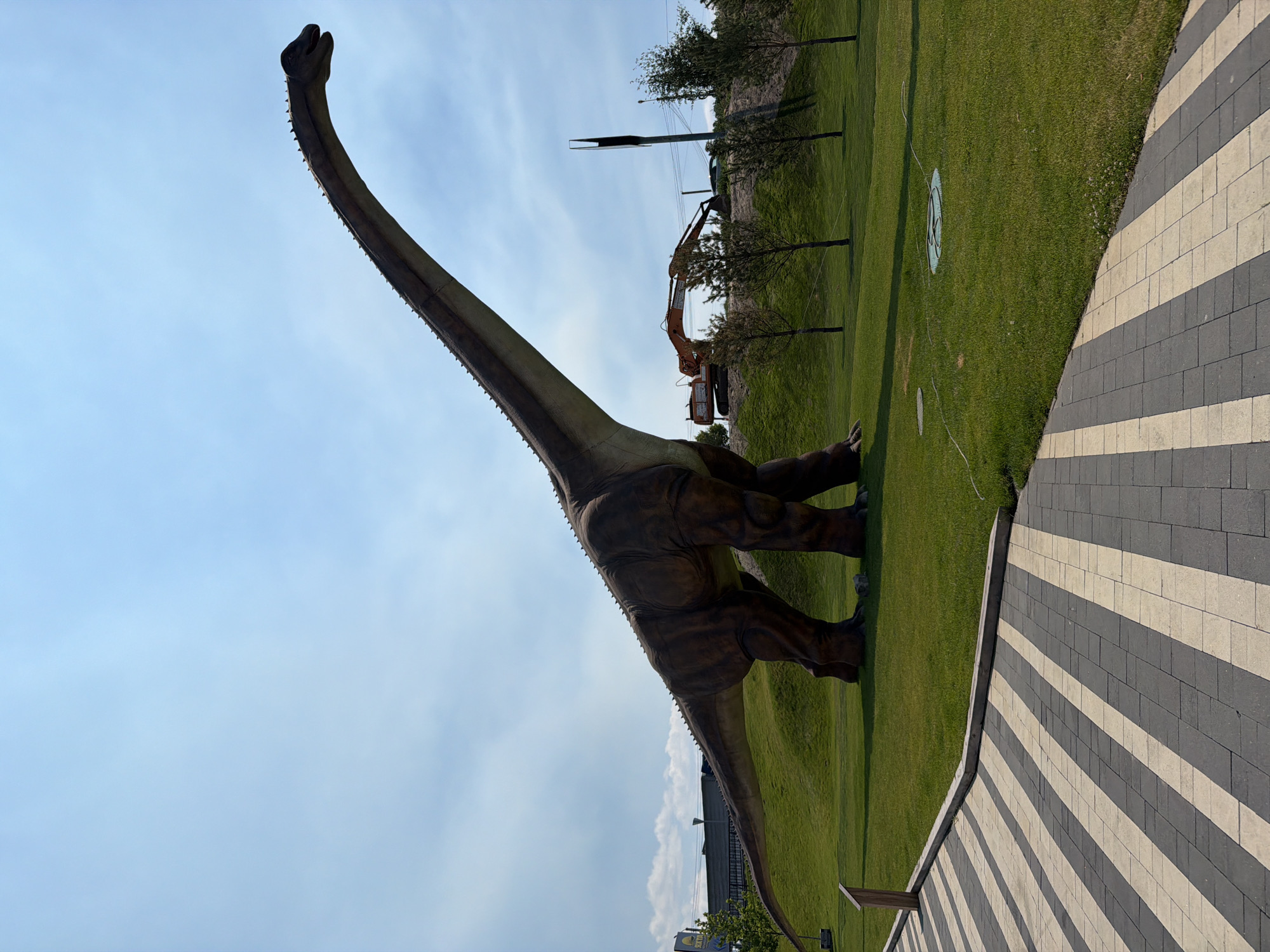 The image shows a large, brown dinosaur statue with a long neck and tail, positioned on a grassy hill. The statue is a prominent subject, taking up a significant portion of the frame. In the background, an orange excavator is visible on a dirt mound, suggesting some sort of construction or land management activity. Several small trees are scattered across the landscape. The ground is covered in lush green grass, and a pathway with alternating dark grey and beige rectangular paving stones is visible in the foreground, bordering the grassy area. The sky is clear blue with some wispy clouds. The overall scene appears to be outdoors in a park-like setting. The lighting suggests it is daytime, likely sunny, given the shadows cast on the grass. The orientation of the image is such that the dinosaur is angled diagonally from the bottom left towards the top right. No people are visible in the frame. No text is discernible in the image. The setting is Khotianivka, Ukraine.