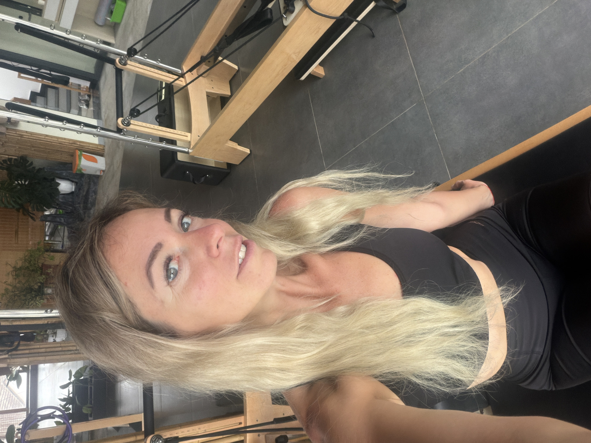 A blonde woman with blue eyes is taking a selfie in a Pilates studio. She is wearing a black sports bra and black leggings. The studio has large windows, bamboo accents, and Pilates reformers. The floor is tiled in gray. The woman is smiling at the camera. The location is identified as Da Lat, Vietnam.