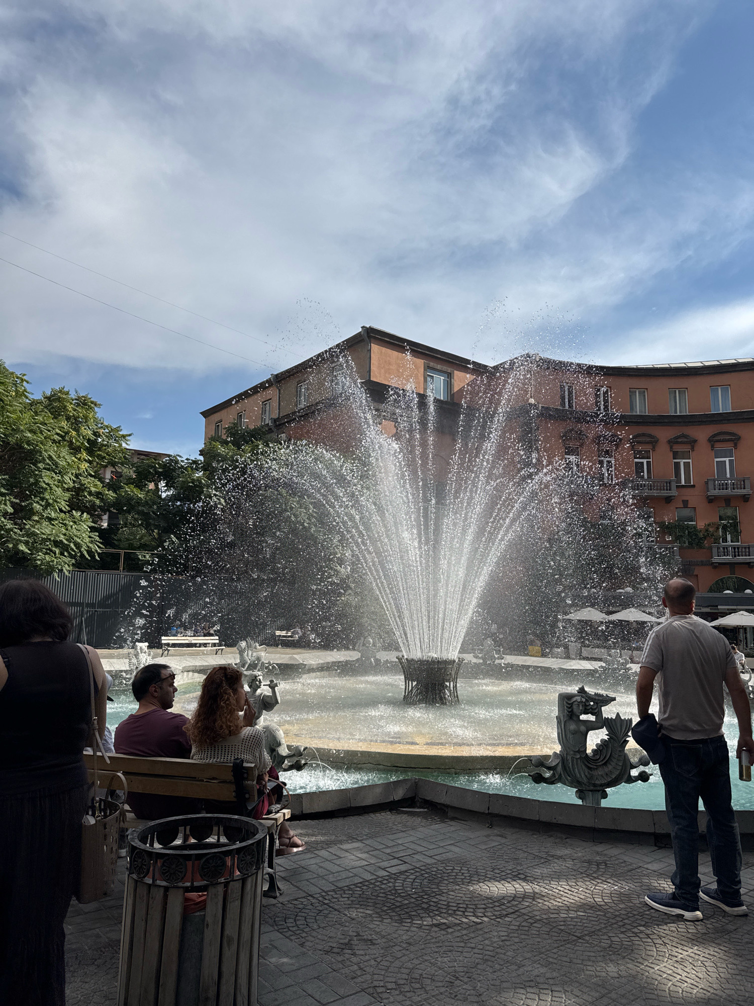 This is an eye-level, medium shot of a public fountain in Yerevan, Armenia, likely located in a prominent city square given the architectural style of the surrounding buildings. The fountain is the central focus, with water spraying outwards in a wide arc. In the foreground, a woman stands with her back to the camera, wearing a black sleeveless dress and carrying a purse over her left shoulder. To her right, a couple sits on a wooden bench; the man, wearing a dark red t-shirt and sunglasses, looks to his left, while the woman, with curly red hair and a white patterned top, looks forward. A large, weathered trash receptacle is positioned in front of the bench.
To the right of the fountain, a man in a grey t-shirt and blue jeans stands facing away from the camera, holding a can in his right hand and a dark jacket or bag in his left. A sculpted mermaid-like figure with a fish tail and intricate detailing is visible on the edge of the fountain. The background features a large, rust-colored, multi-story building with arched windows and balconies, characteristic of Soviet-era architecture often found in Yerevan. Lush green trees are visible to the left of the building, and a few white umbrellas suggest outdoor seating for a cafe or restaurant behind the fountain. The sky is a bright blue with scattered white clouds, indicating daytime. The ground is paved with patterned tiles.