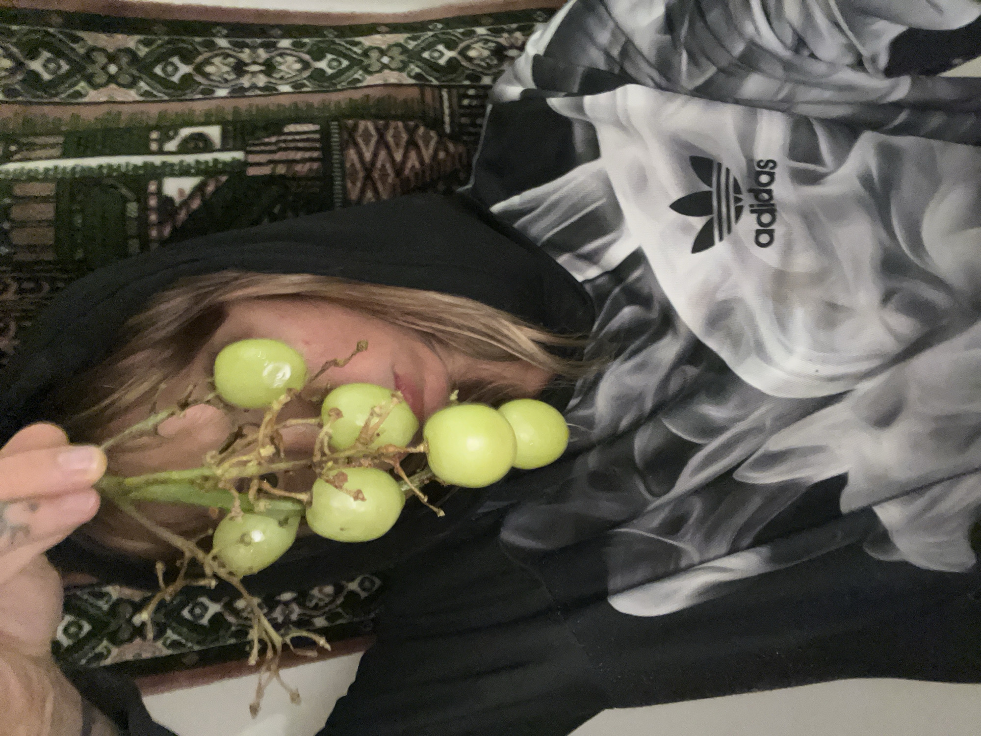 A person wearing a black hoodie lies on a patterned surface, possibly a bed or couch, holding a cluster of green grapes in front of their face. An Adidas logo is visible on a black and white patterned fabric next to the person. The background is slightly blurred. There is no information in the image to indicate the location is Buenos Aires, Argentina.