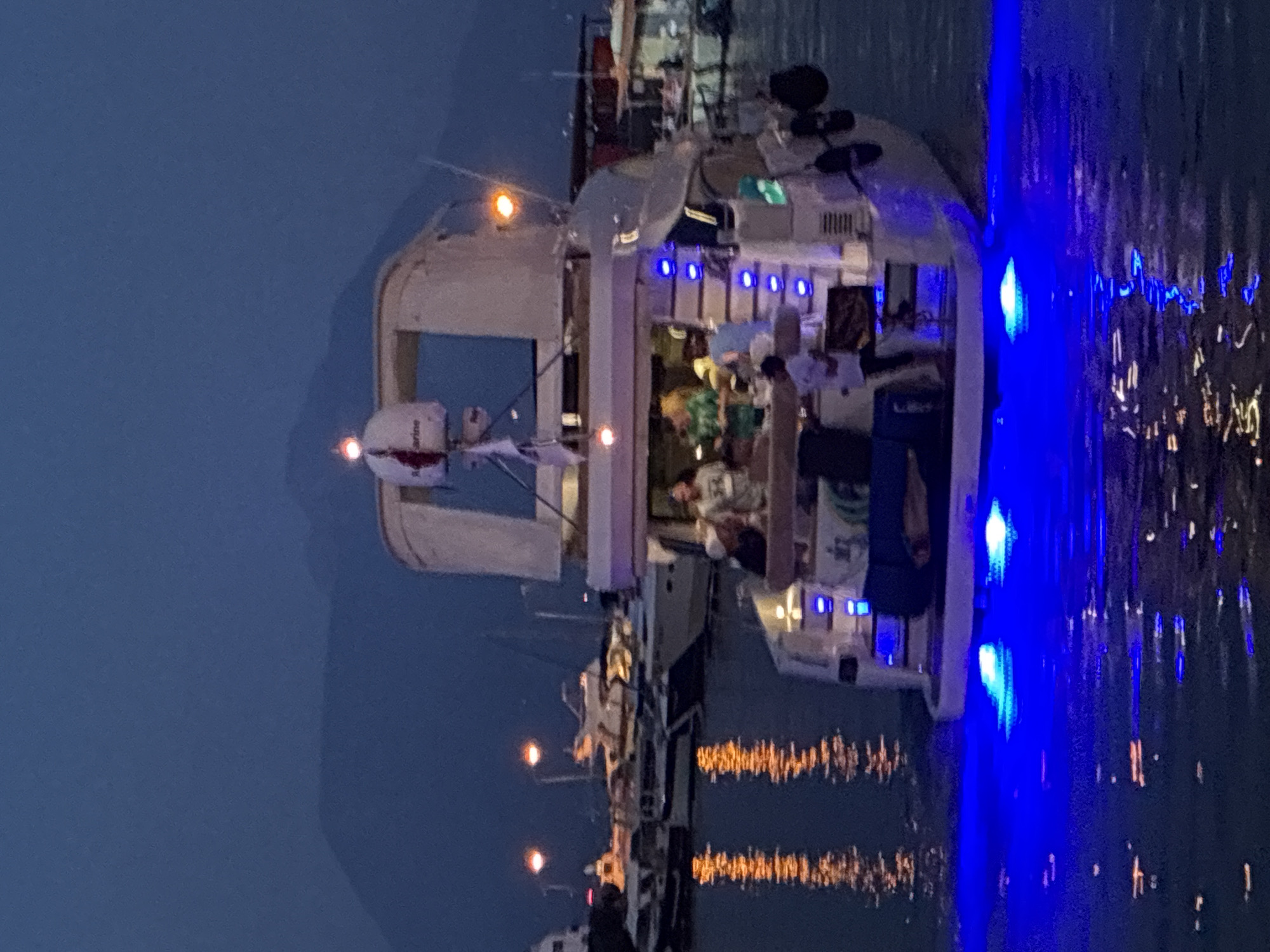 The image depicts a night scene at a harbor in Alanya, Turkey. A yacht is the central focus, illuminated by blue underwater lights that cast a glow on the water. Several individuals are visible on the yacht's deck, engaged in what appears to be a social gathering. In the background, other boats are docked, and distant lights suggest the presence of a town or city. Mountains are faintly visible in the far background against the twilight sky. The overall atmosphere is one of a calm evening by the sea.
