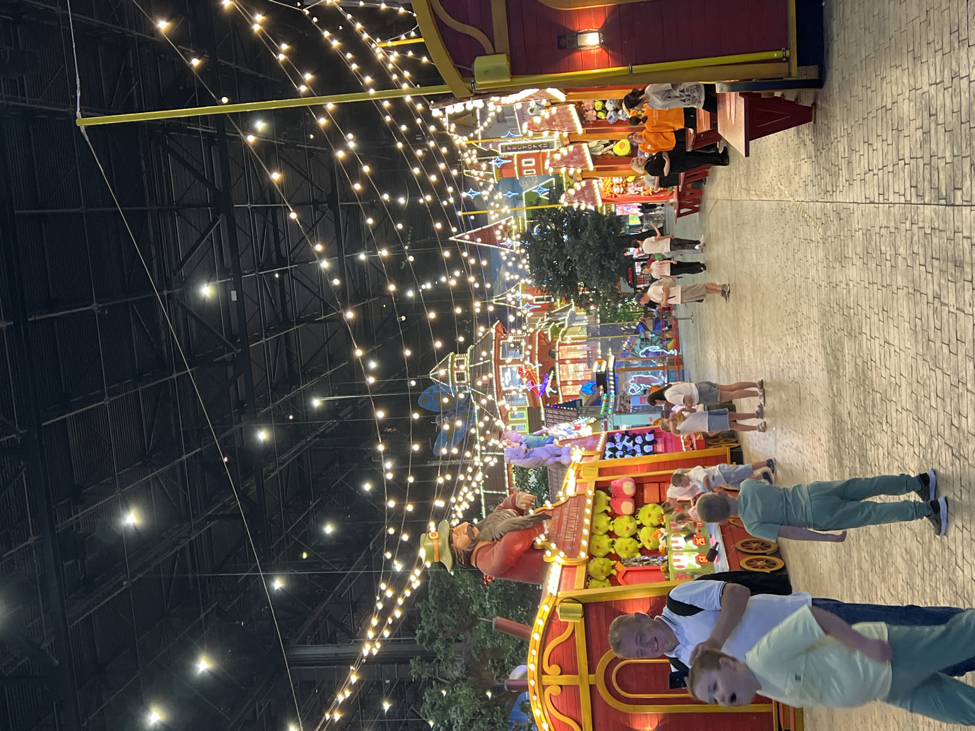 The image depicts a lively indoor amusement park scene in Moscow, Russia, likely at Ostrov Mechty (Dream Island). A pathway paved with interlocking bricks leads through a brightly decorated area, illuminated by numerous strings of warm-toned fairy lights suspended from the high ceiling. The architecture resembles a whimsical European-style town with colorful buildings, towers, and playful embellishments.
In the foreground, a man in a white polo shirt and blue jeans, holding a smartphone, walks with a young boy in a light green t-shirt and matching pants. The boy is looking to his left, mouth open in what appears to be a yell or excited exclamation. To the right of them, another boy in a similar light green outfit stands facing away from the camera, looking towards the amusement stalls.
Further down the pathway, several people are strolling and interacting. On the left, a red amusement stall with a large, animated figure of a man in a green hat and red jacket is visible, holding a grey object. The stall is adorned with rows of yellow and pink plush toys. To the right, more amusement stalls and colorful structures are present, with people gathered around them.
The environment suggests a recreational setting designed for entertainment, with vibrant colors, decorative lighting, and attractions characteristic of an amusement park. The overall impression is one of activity and festivity.