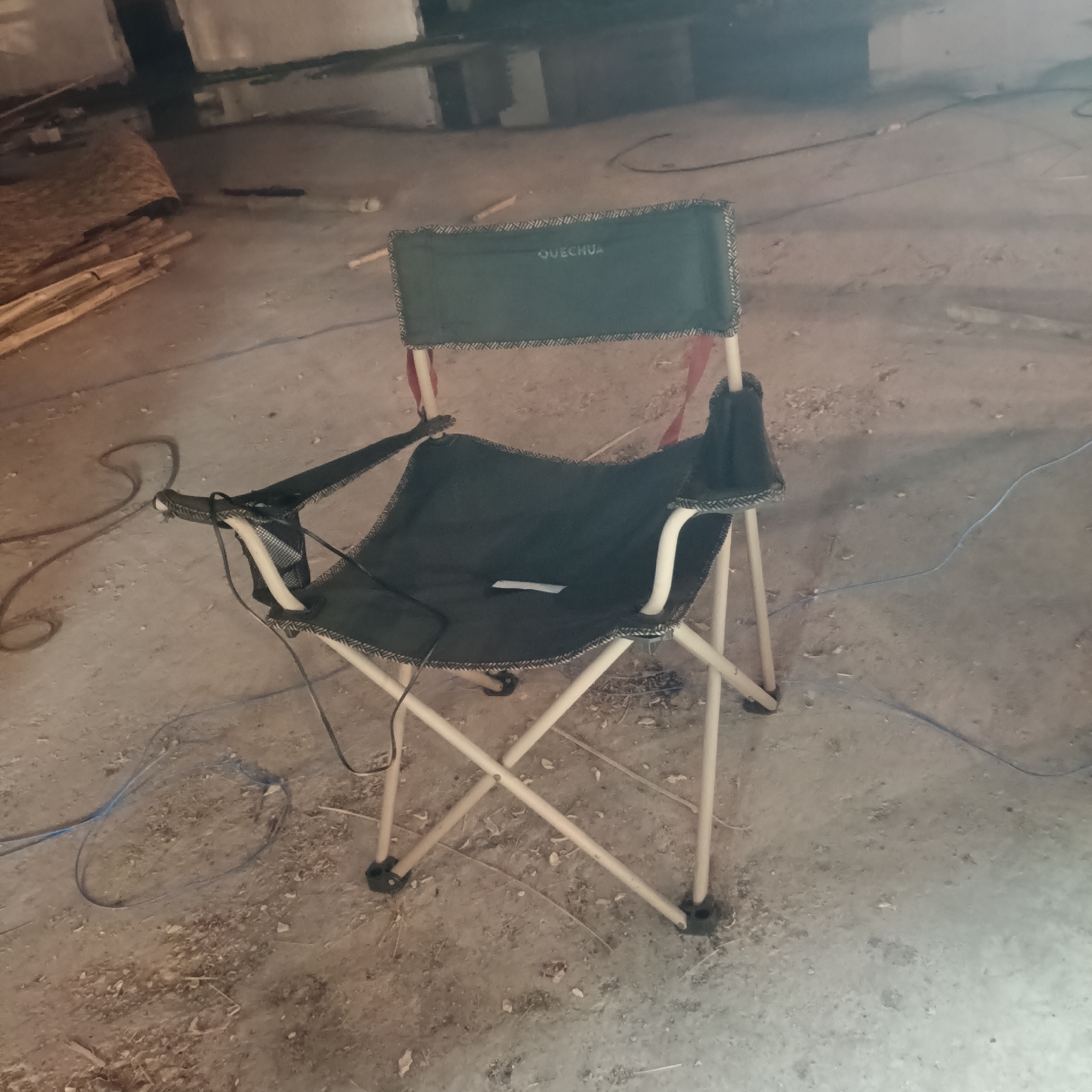 A green Quechua camping chair with armrests stands on a concrete floor. The area appears to be a storage space or basement, with lumber and other objects visible in the background. Blue and black wires are present on the floor around the chair. The location is unspecified within Panaji, India.