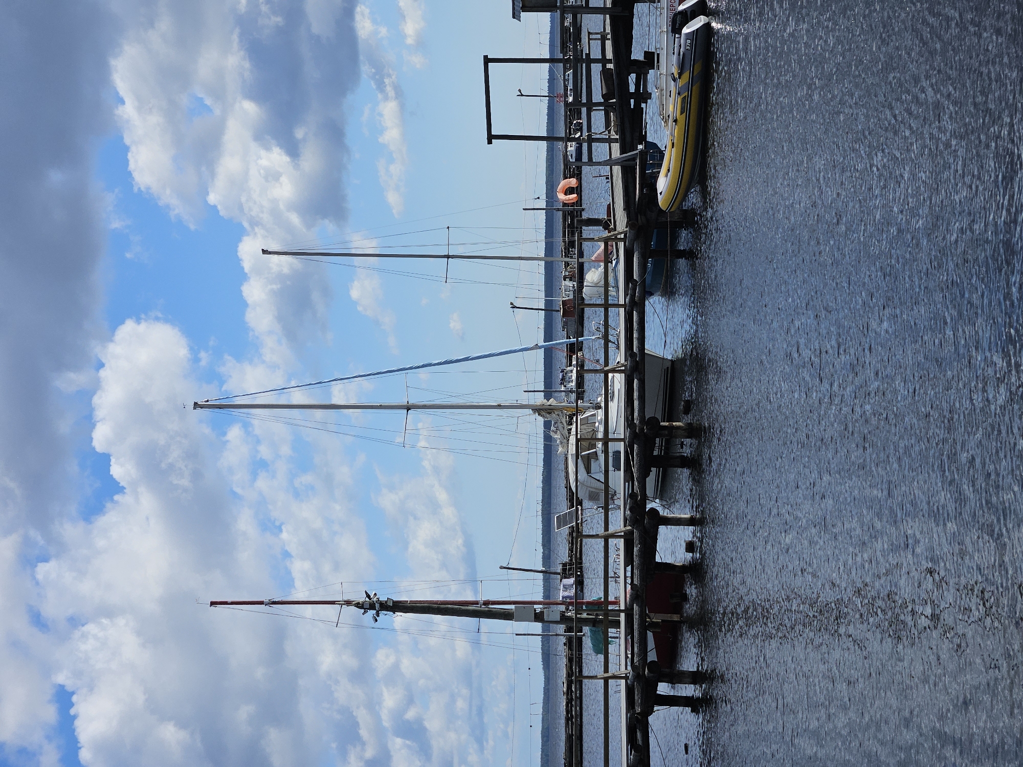 A wide shot shows a calm body of water with a wooden pier extending into it. Several sailboats are docked at the pier, with masts reaching towards a bright blue sky filled with fluffy white and gray clouds. On the right side of the pier, a yellow and gray inflatable boat with an outboard motor is moored. The background shows a distant treeline under a hazy horizon. The water reflects the sky and the pier structure. There are no people visible in the image.