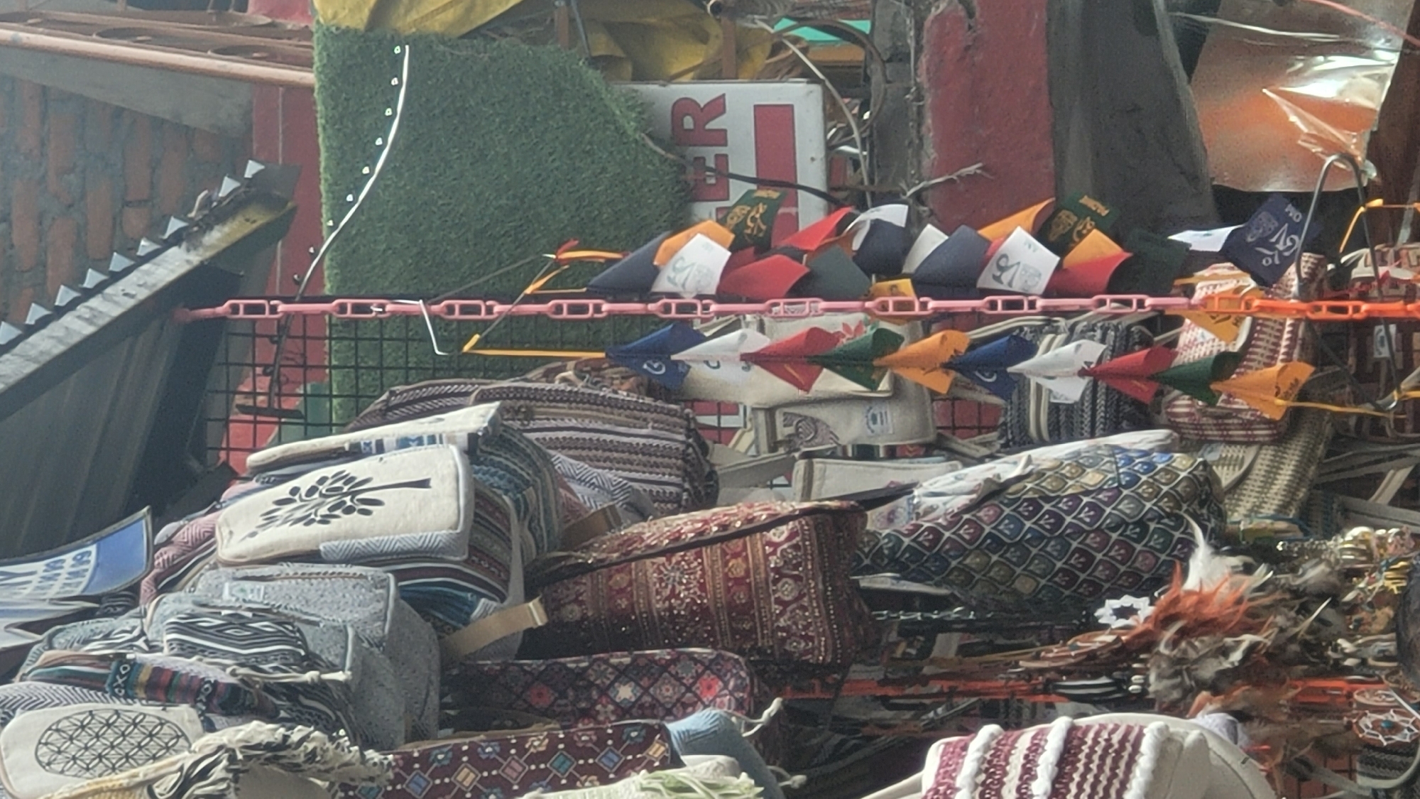 The image depicts a market stall in Vashisht, India, laden with a variety of textiles and decorative items. Prominently displayed are numerous bags of different sizes and designs. Some are made of woven fabric with intricate patterns, while others appear to be canvas or embroidered. Several bags feature ethnic prints, including floral motifs and geometric shapes. Hanging from a pink plastic chain are small prayer flags in various colors, such as red, blue, yellow, green, and white, many of which bear Buddhist symbols or inscriptions. A large green artificial turf panel is visible in the background, along with a sign with red lettering and the partial word "ER." The overall scene suggests a vibrant and cluttered marketplace setting.