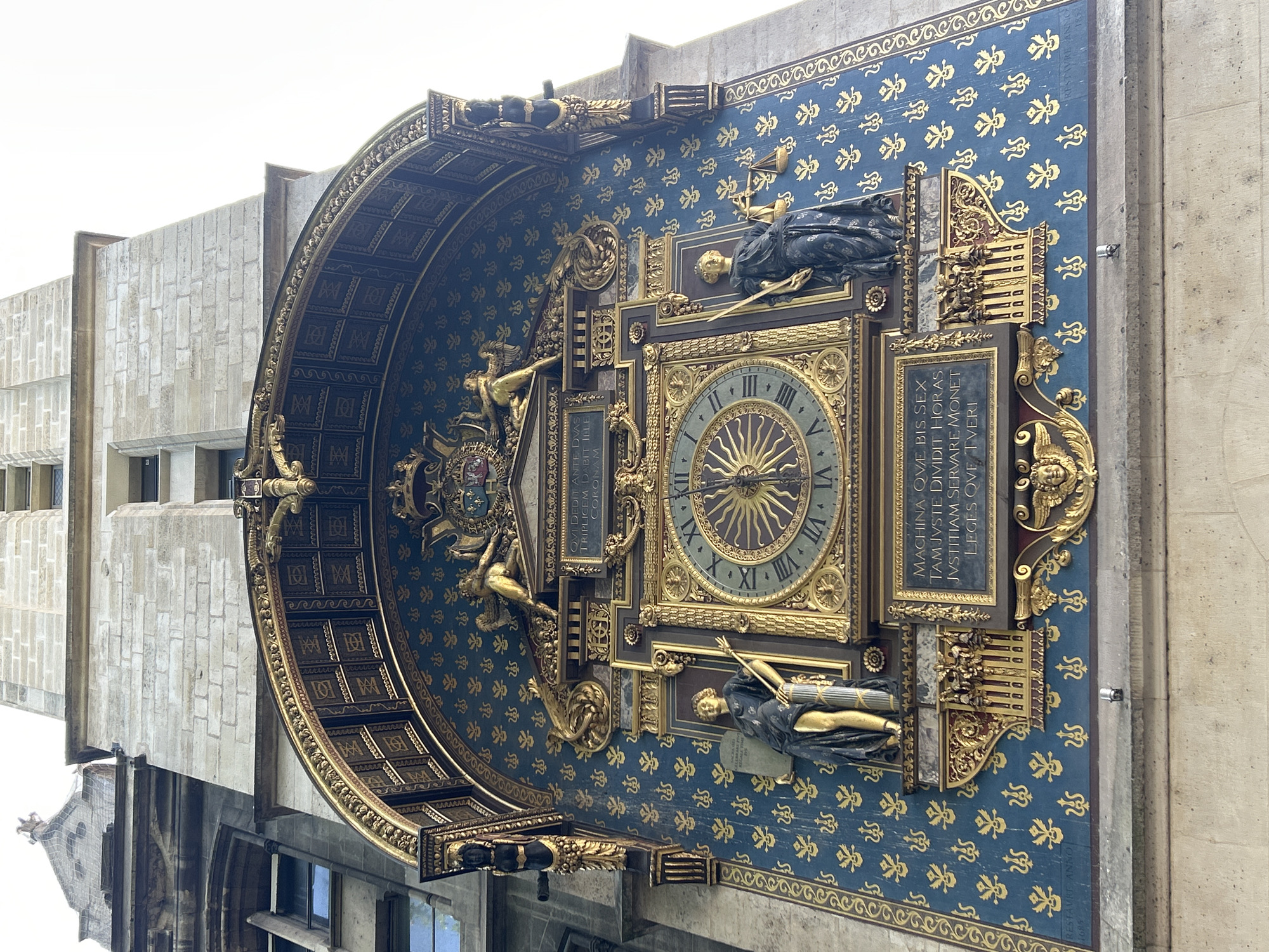 The image depicts the Astronomical Clock (L'horloge astronomique) located in the Conciergerie building, Paris, France. The clock face is ornate, with Roman numerals and a sun-shaped hand. Flanking the clock are two allegorical statues: one holding a tablet and a scepter, and the other holding a scale. The clock is set against a deep blue background adorned with gold fleur-de-lis motifs. Above the clock, there are gilded figures of angels and a crest. The clock is framed by a decorative arch and gold detailing. The structure is made of stone, with a white sky visible at the top. The inscription "MACHINE OVE BIS SEX TANT IVSTE DIVIDI HORAS IVSTITHAM SERVARE MONET OVE TVERI" is visible below the clock. Another inscription at the bottom left reads "RESTAVRE. ANNO. 1685".