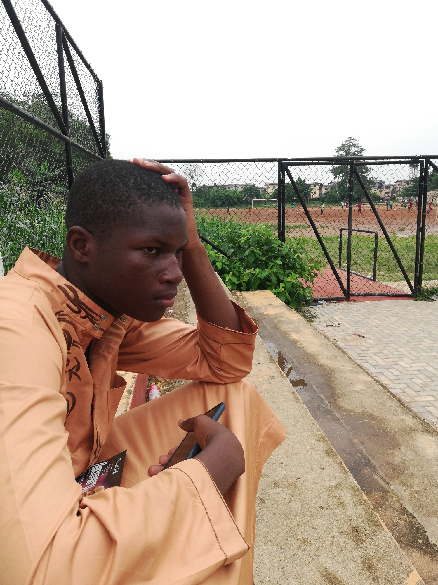 A medium shot captures a young man sitting on a concrete ledge outdoors. He is dressed in a light orange traditional garment with intricate designs on the sleeves and chest. The man's left hand rests on his head, suggesting a pensive or thoughtful mood. In his right hand, he holds a dark-colored smartphone. He is positioned in front of a chain-link fence that separates him from a sports field. On the field, there are people engaged in a soccer game. The area appears to be a recreational space, possibly a park or schoolyard. The sky is overcast, indicating a cloudy day. There is no mention of the location in the city of Unknown, Unknown, in the image.
