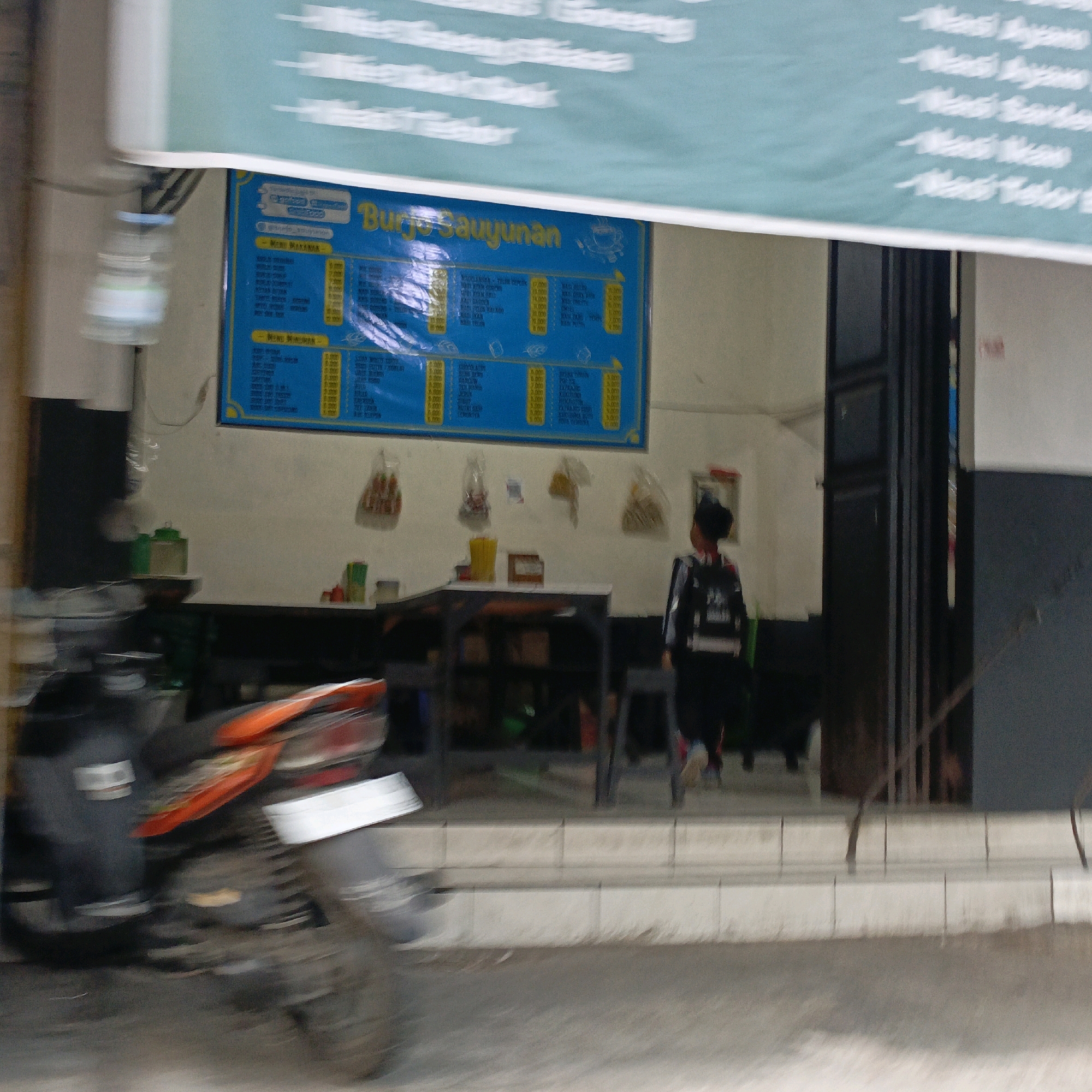 In Salatiga, Indonesia, a child wearing a dark jacket with white stripes walks past a food stall. The stall has a blue menu board displaying various food and drink items, including "Nasi Ayam" and "Nasi Ikan." Bags of snacks are hung on the wall behind the counter. In the foreground, a motorcycle is parked, with its red and orange parts blurred due to motion. The overall scene suggests a busy street setting with a focus on food service.
