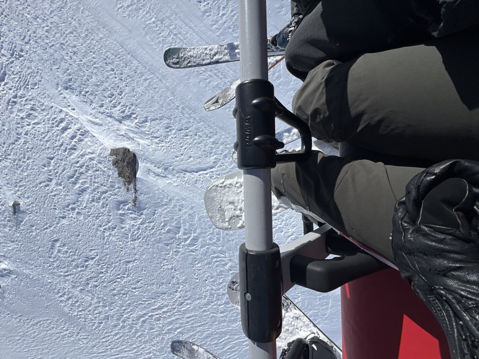 The image captures a close-up view from within a ski lift cabin, looking down towards the snowy slope below. The cabin is occupied by a single individual, whose lower legs and feet are visible. Their right leg is extended, clad in dark grey ski pants, and their left leg is bent at the knee, with their foot resting on the lift's metal bar. They are wearing a pair of black ski gloves, one of which is visible on their left hand. The scene is set against a backdrop of a snow-covered slope, with the texture of the freshly fallen snow clearly visible. The coordinates provided (latitude: 45.421, longitude: 7.013) pinpoint the location as Val-d'Isère, France. There are two pairs of skis secured to the lift's frame, indicating the individual is a skier. The image was likely captured during a sunny winter day as the bright white snow reflects the light. The overall atmosphere is one of tranquility and quietude, typical of a winter ski resort.