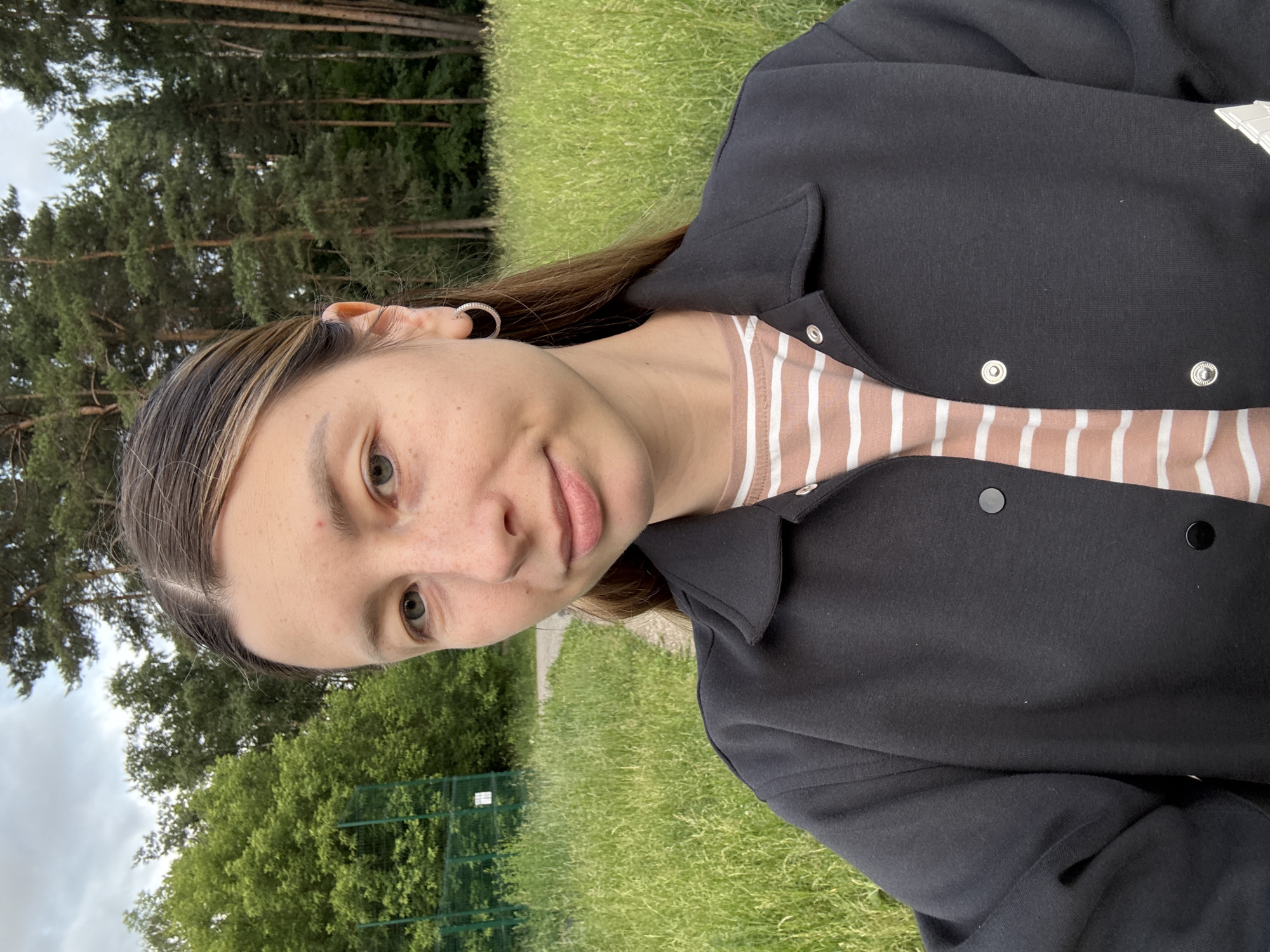 A young woman with light brown hair, fair skin, and freckles is taking a selfie outdoors. She is wearing a black jacket over a striped t-shirt. Her hair is pulled back, and she is wearing a small hoop earring in her left ear. She has a slight smile. The background shows a grassy field and a line of trees. A green fence and a white sign are visible on the left side of the image, suggesting a park or sports field. The sky is overcast. The scene is likely in a park in Moscow, Russia, given the type of trees and the overcast sky, common in the Moscow region during certain times of the year.