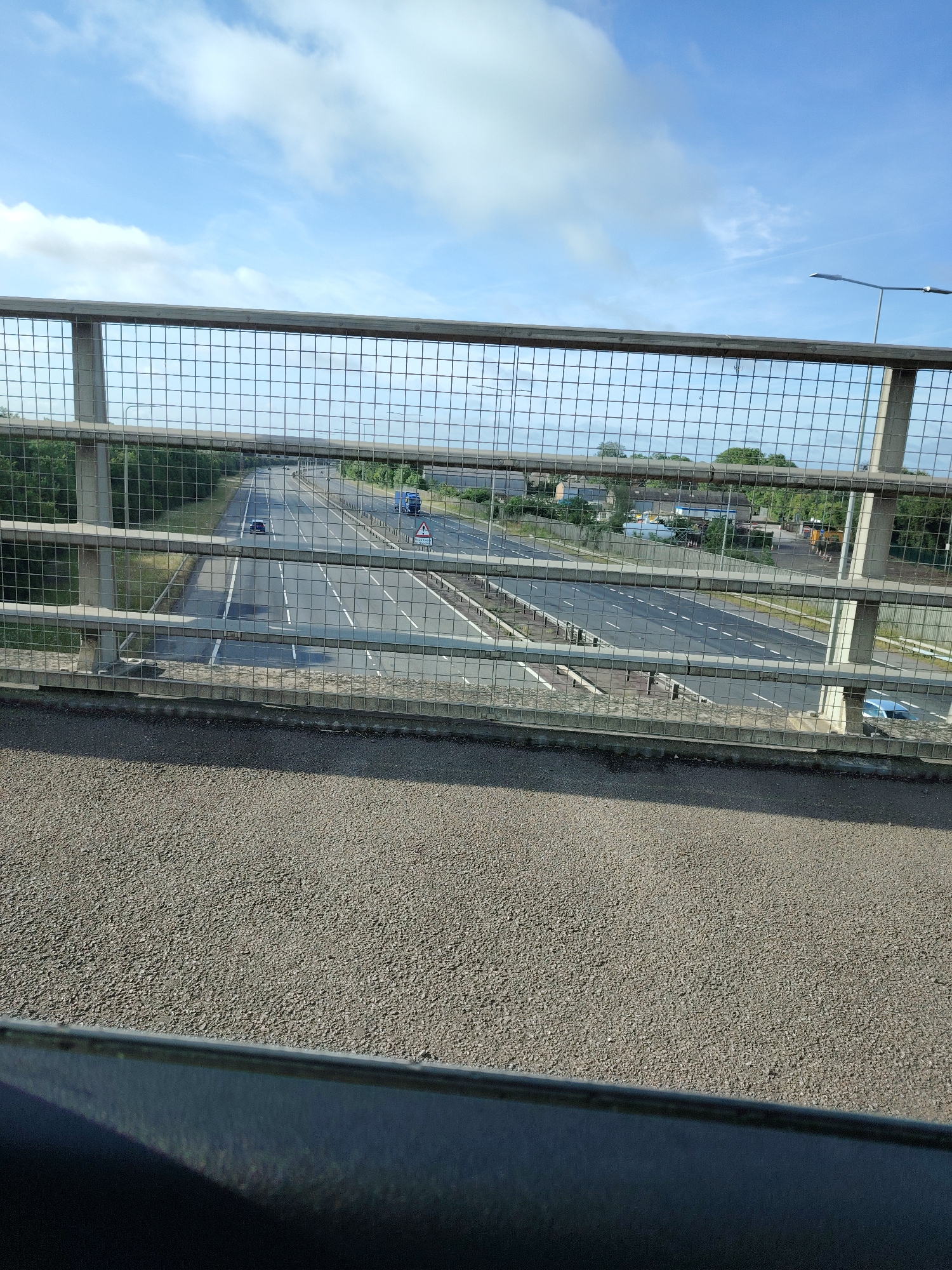 The image presents a forensic view from an elevated bridge overlooking a multi-lane highway in Huntingdonshire, United Kingdom.
In the immediate foreground, a dark, indistinct horizontal surface occupies the bottom portion of the frame, surmounted by a light grey, granular asphalt surface characteristic of a pedestrian walkway. Above this, a prominent metal mesh fence with a grid pattern acts as a barrier, partially obscuring the view of the highway. The fence features horizontal and vertical support bars.
Through the mesh fence, a dual-carriageway highway, consistent with a UK motorway or major A-road, is visible. Each carriageway features at least three lanes, delineated by white road markings including solid lines, dashed lines, and arrows on the dark grey asphalt. A central reservation, containing some sparse vegetation, separates the two directions of traffic.
On the left carriageway, which appears to lead away from the viewer, a dark-colored car is positioned in a middle lane. On the right carriageway, potentially carrying traffic towards the viewer, a blue articulated lorry (truck) is present in a middle lane, while a smaller light blue car is partially visible in the far-right lane near the bottom right corner of the highway view. A triangular warning sign with a red border, commonly indicating roadworks or a temporary hazard, stands on the central reservation or hard shoulder of the right carriageway.
Supporting infrastructure includes large, grey concrete pillars, which are part of either the bridge the photograph is taken from or another overhead structure. A tall streetlight with a horizontal arm extends from the right side of the highway.
The background features dense green trees lining both sides of the highway, extending into the distance. Beyond the trees on the right, several light-colored buildings with grey roofs, indicative of an industrial or commercial area, are visible. The sky above is predominantly clear and light blue, with scattered, patchy white cumulus clouds, suggesting bright daylight conditions. There are no identifiable people or explicit actions captured in the scene. The overall environment is consistent with a transportation artery in a semi-rural or exurban area of Huntingdonshire, likely a section of the A1(M) or A14.