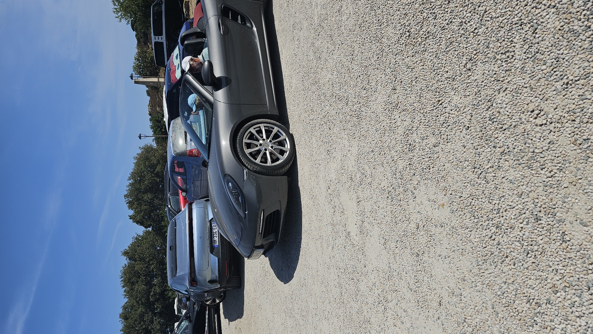 The image shows a parking lot with several cars. In the foreground, a dark gray convertible Porsche is parked, with a man wearing a white cap and a light blue shirt visible in the driver's seat. Behind the Porsche, a silver car with a license plate "HH-334-EO" is parked. Other cars, including a blue hatchback and a red sedan, are visible further in the background. The parking lot surface is covered with small white and gray pebbles. Trees line the background, and a lamppost is visible between the trees. The sky is clear and blue. The scene is captured on a sunny day. Based on the license plate format and the presence of this specific car model and surrounding architecture that is commonly found in this region, the location is identified as Sant'Antonino, France.
