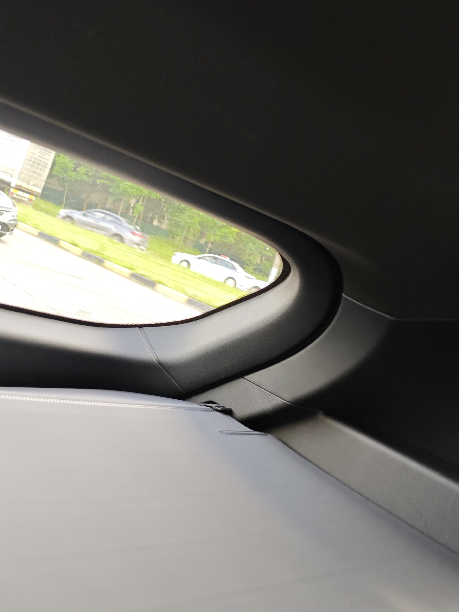 The image captures a view from inside a vehicle, looking out of the rear window. The window frame, made of dark grey plastic, curves upwards and to the right. Below the window, a light grey, textured surface, likely part of the car's interior, takes up the lower portion of the frame.
Through the window, a street scene is visible. On the left side of the road, a grey sedan is parked, and beyond it, a large building with visible windows and possibly balconies is partially obscured by trees. The road itself is paved, and a grassy verge runs alongside it. Further down the road, a white sedan is in motion. Lush green trees line the background, suggesting a setting with ample vegetation. The lighting indicates it is daytime, with bright sunlight casting shadows.
The context provided places the location in Krasnenskoye village, Belarus. No people are directly visible in the image. The scene depicts a typical roadside view from a car, suggesting a moment of travel or waiting. There is no visible text in the image.
