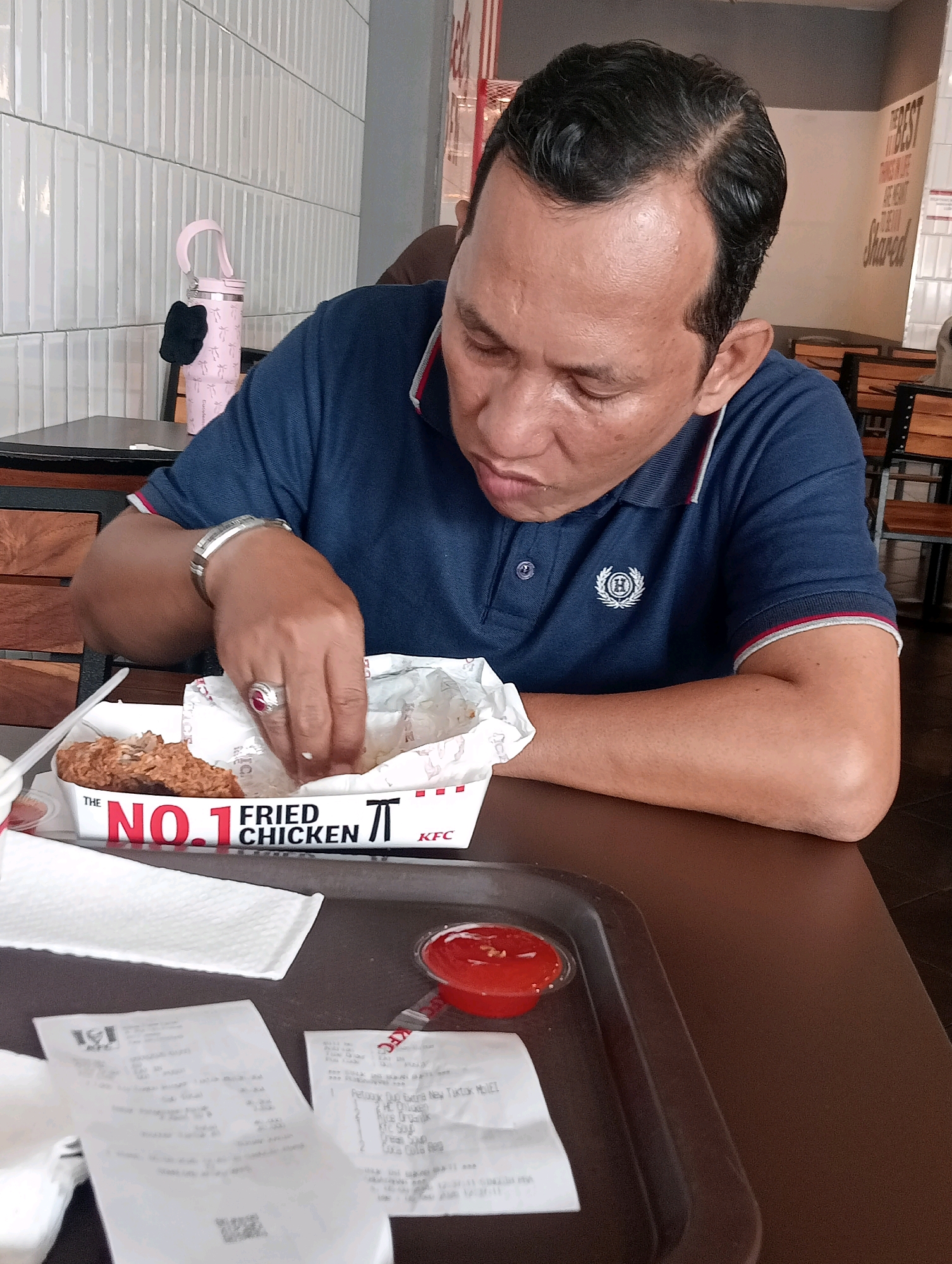 A man is eating fried chicken at a KFC restaurant in Jambi, Indonesia. The man is wearing a blue polo shirt and is sitting at a brown table. He is holding a piece of fried chicken in his hand and is about to eat it. There is a tray with ketchup and receipts on the table. The restaurant has white tiled walls and wooden chairs. A pink water bottle with a black keychain is on a table behind the man.