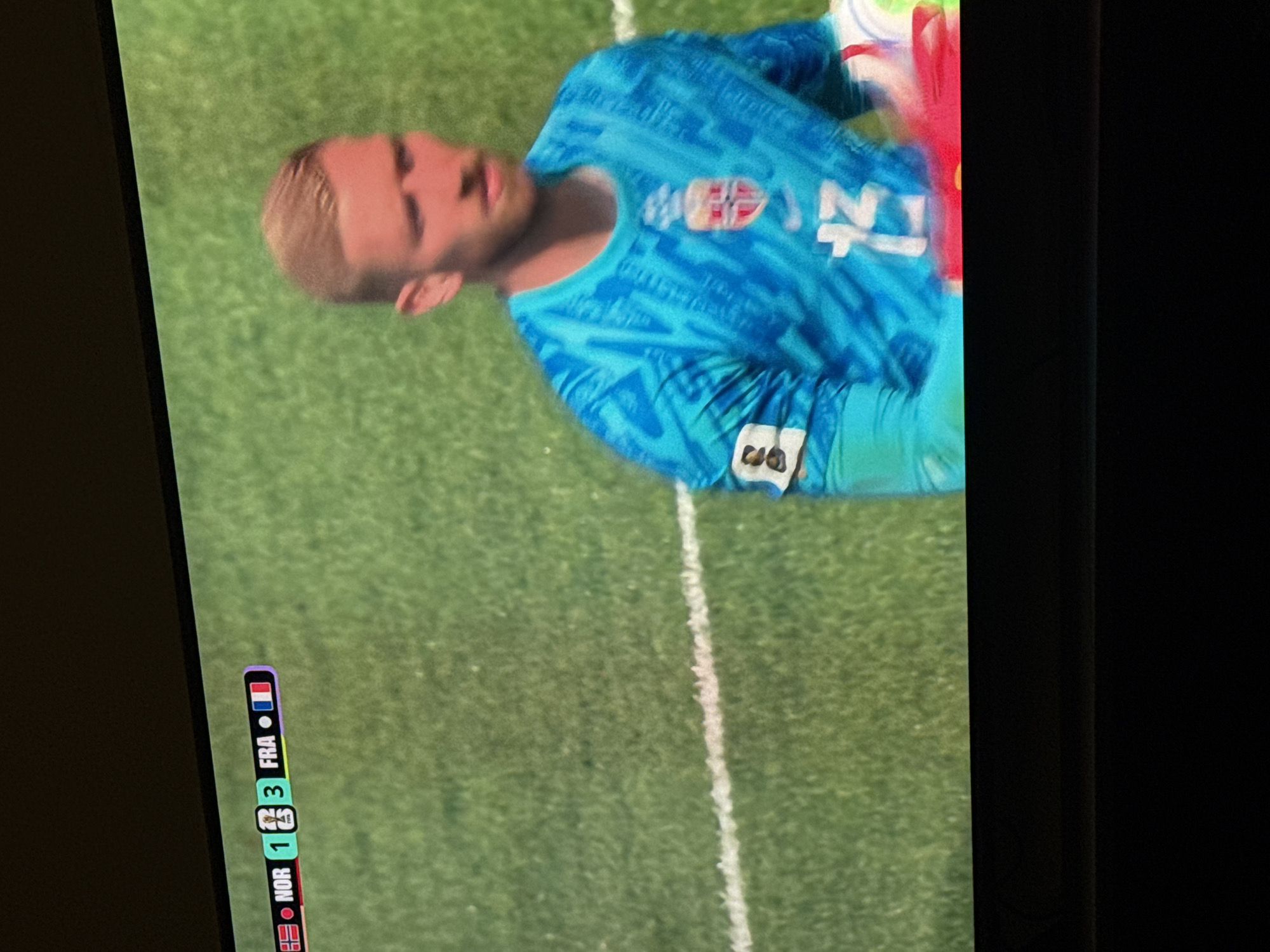 The media file shows a close-up, vertical shot of a young male soccer player wearing a blue jersey with a white pattern and a crest on the left chest. He has blonde hair and appears to be a goalkeeper, with some blurring suggesting motion. The player is looking down and to his right, with a neutral expression. He is standing on a green soccer field with a white line visible to his right. In the lower portion of the image, a score graphic is displayed vertically, showing "NOR 1 3 FRA" with small flags representing Norway and France. The context provided indicates the location is Moscow, Russia, though this is not visually evident in the image itself. The lighting suggests daytime. The overall impression is a moment captured during a soccer match.