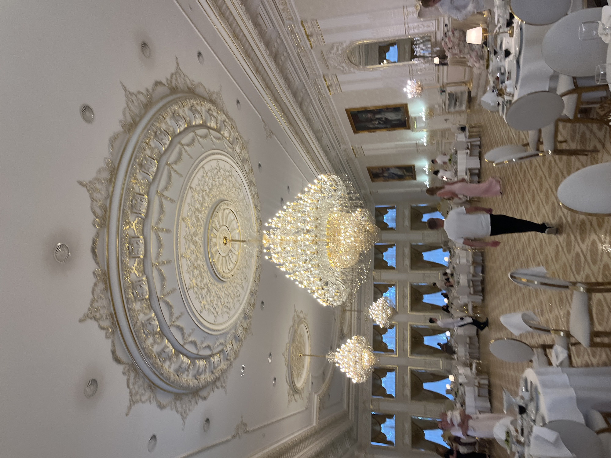 The media file depicts a grand indoor event, possibly a banquet or wedding reception, set in a lavishly decorated hall. The ceiling is a prominent feature, adorned with intricate white and gold plasterwork, including a large circular medallion with ornate detailing and smaller decorative elements. Several large, sparkling crystal chandeliers hang from the ceiling, illuminating the scene. Paintings in ornate frames are visible on the walls, suggesting a formal setting.

The hall is set up with numerous round tables covered in white tablecloths, each meticulously arranged with place settings. White chairs with gold accents surround the tables. Several people are visible in the image, some dressed in white uniforms, likely serving staff, and others in formal attire. One person in a long pink dress is walking through the hall. The overall impression is one of elegance and celebration.

The lighting suggests it is likely evening or indoors without natural light being the primary source. The location cues are minimal, but the description of ornate decor and grand chandeliers points to a high-end venue. No visible text is present in the image.