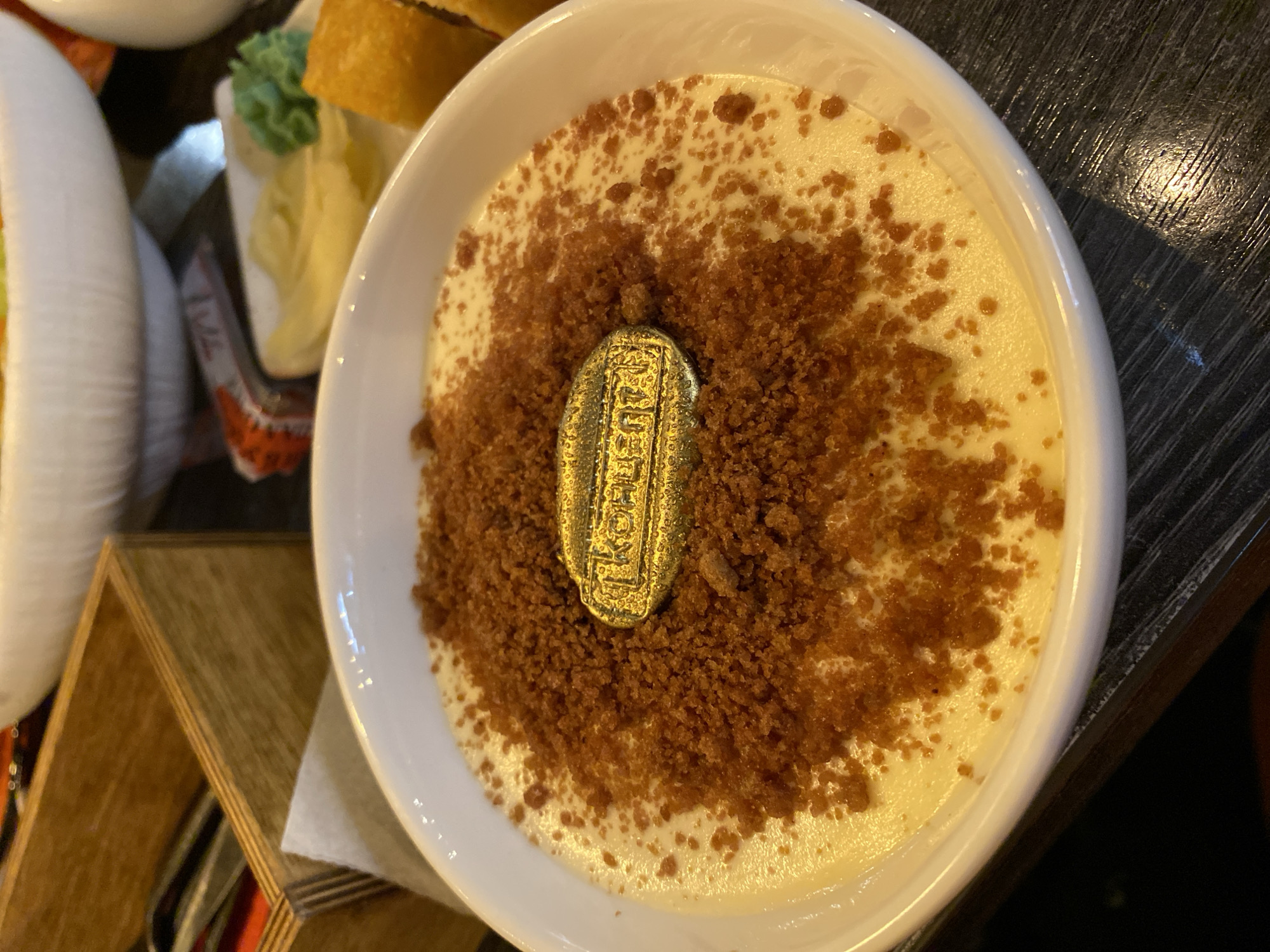 A close-up shot depicts a dessert in a white bowl. The dessert has a creamy, light-yellow base sprinkled with brown crumbs and features a gold-colored, rectangular object in the center, possibly a decorative piece. The bowl is placed on a wooden surface with a slightly glossy finish. Additional dishes are partially visible to the left of the bowl, including what appears to be a white cylindrical object and a dish containing yellow and green ingredients. The location is not provided in the image.
