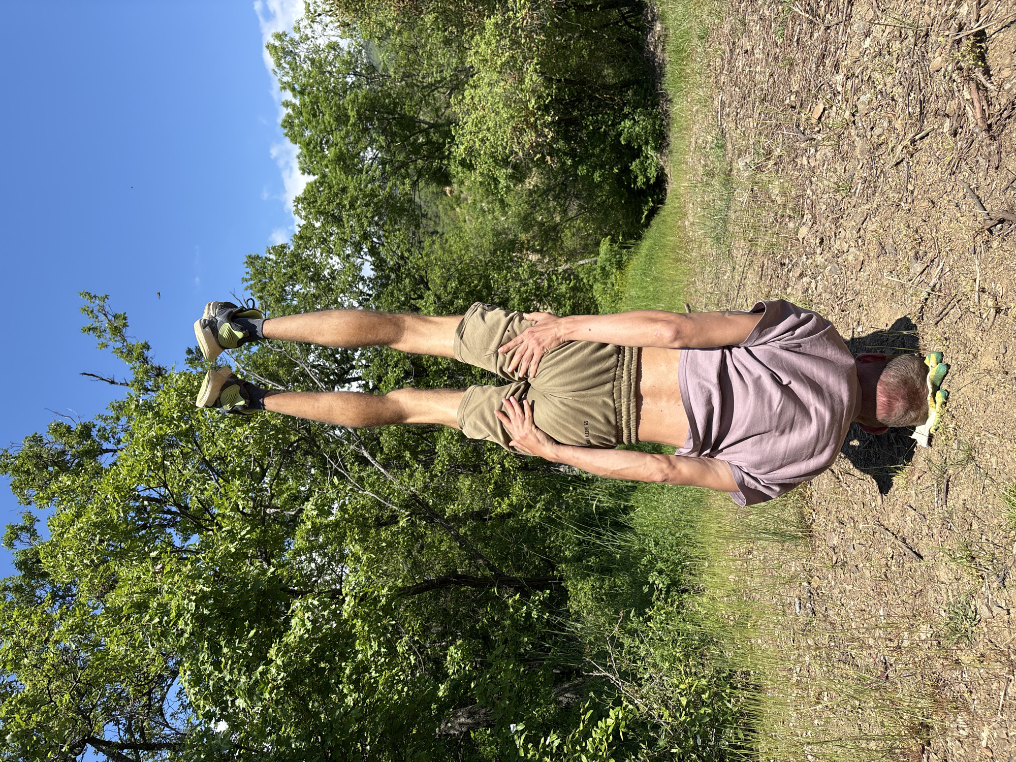 A man is performing a handstand outdoors. He is wearing a light purple t-shirt, khaki shorts, and athletic shoes. His legs are straight and pointing towards the sky. He is balancing on his hands on a patch of dirt and grass. In the background, there are trees and a blue sky with some clouds. This image does not contain enough information to confirm the location in the city of Nizhnee Zaprudnoe, Ukraine.