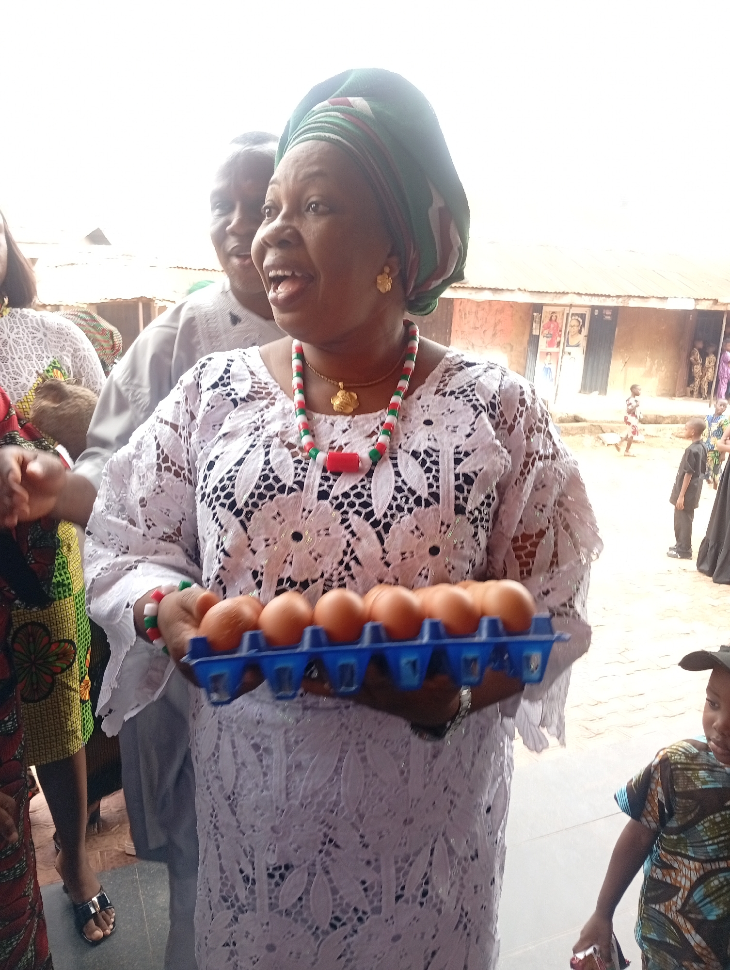A woman in Narayi, Nigeria, wearing a white lace dress, a green and white head wrap, and a necklace of green, red, and white beads, holds a blue egg carton filled with brown eggs. A man stands behind her to her left, and other people are partially visible on either side of her. A building with a tin roof is in the background. Children are visible standing outside the building.