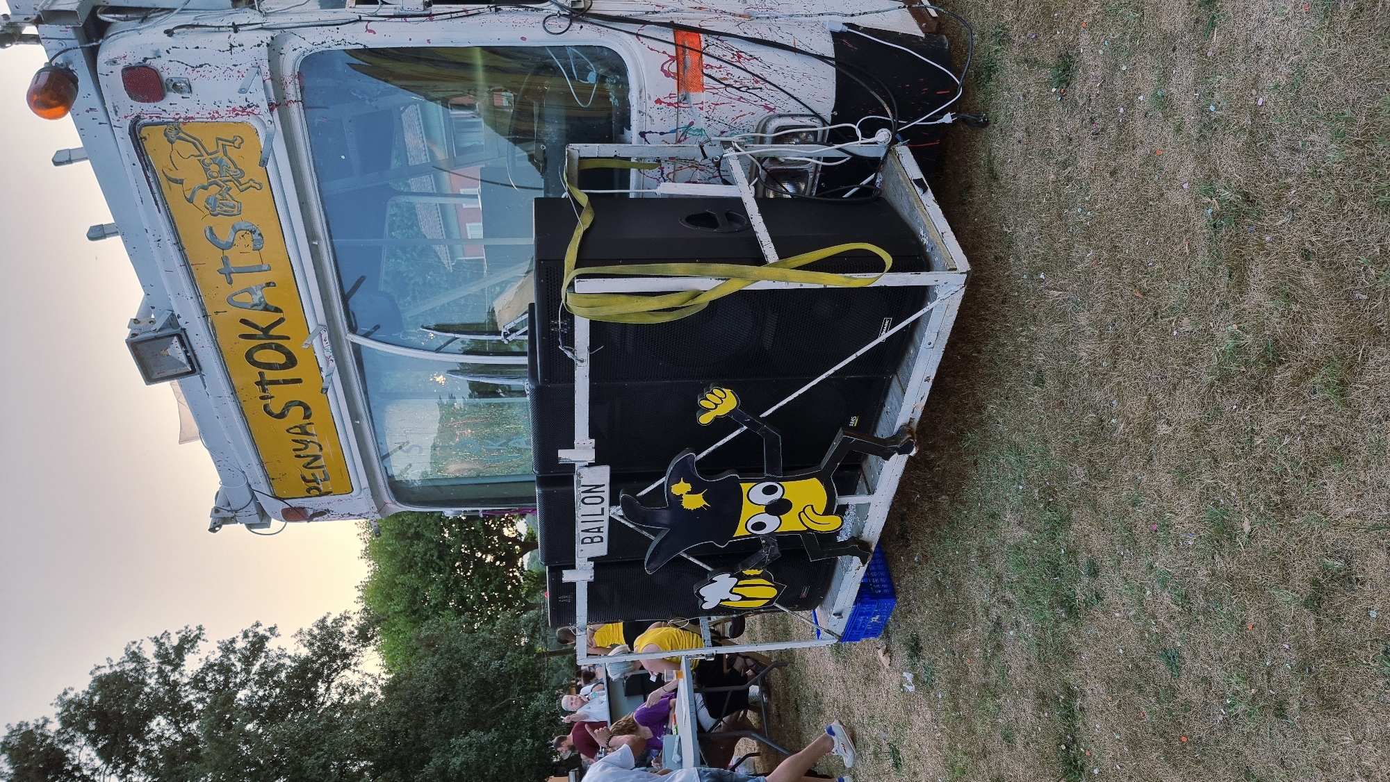 The image shows a mobile sound system mounted on a trailer, likely used for outdoor events. The trailer is white with red and pink paint splatters. At the front of the trailer, a yellow character with black outlines is depicted, holding a mug of beer and wearing a pointed hat. The character is positioned in front of large black speakers. A sign above the speakers reads "BAILON". A yellow strap is visible, securing some of the equipment. Above the trailer, a yellow sign with red lettering reads "PENYAS'TOKATS" and features a drawing of two figures. In the background, trees and a partially visible building are present. Several people are seen gathered around tables in the background, suggesting an event is taking place. The ground is covered with dry grass and scattered confetti. The scene appears to be set in an outdoor, grassy area, possibly a park or field. The overall impression is one of a festive, informal gathering.