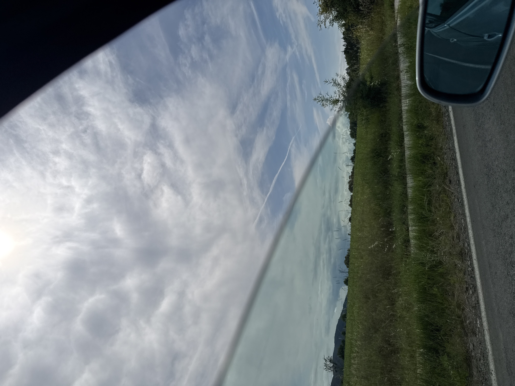 A horizontal cloud band stretches across a bright sky, obscuring the sun in Tsarevo, Bulgaria. Below the cloud band, a vibrant green field extends to a tree line and distant hills under a partly cloudy sky. A road runs in the foreground, with a car's side mirror visible in the lower right corner, reflecting the scene.