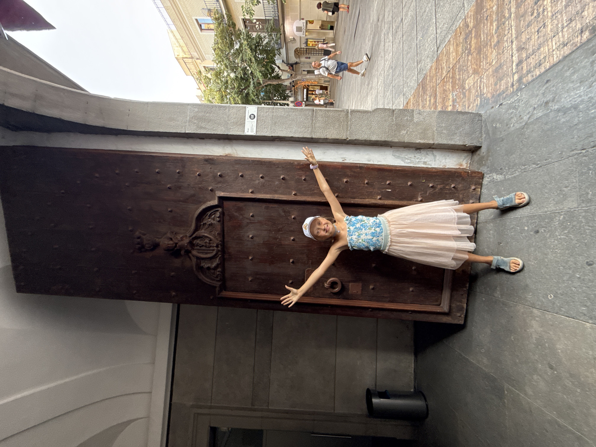 A young girl wearing a blue and white floral top, a light pink skirt, a white cap, and blue sandals stands with her arms outstretched in front of a large, dark wooden door. The girl is positioned slightly to the left of the door, which appears to be part of an old building. The door is ornate, with decorative carvings at the top and numerous dark studs. To the left of the door, a dark grey trash can is visible. The ground is paved with dark grey stone. To the right of the girl and the door, a concrete archway reveals a street scene. A man in a grey t-shirt and blue shorts walks away from the camera on the right side of the street, and other people are visible further down the street in front of shops. The street is paved with a mix of grey and reddish-brown wooden planks. A sign with the number "112" is mounted on the concrete pillar of the archway. The architecture suggests a historical urban setting in Mataró, Spain. The lighting indicates it is daytime, possibly late afternoon.