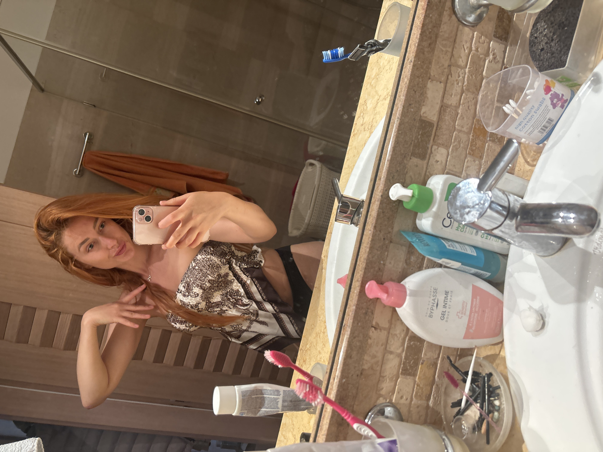 A woman with long, auburn hair is taking a mirror selfie in a bathroom. She is wearing a strapless top with a brown and white pattern and black underwear. She is holding a pink smartphone in her right hand. The bathroom has a marble countertop with various toiletries, including toothbrushes, lotions, and cotton swabs. A towel rack with an orange towel is visible in the background, as well as a shower enclosure. The image is taken at eye level, with the woman slightly angled towards the camera. The lighting is soft, creating a warm and intimate atmosphere. The overall scene suggests a private moment in a personal space, likely during a vacation or trip, possibly in Budva, Montenegro, given the context of the prompt.