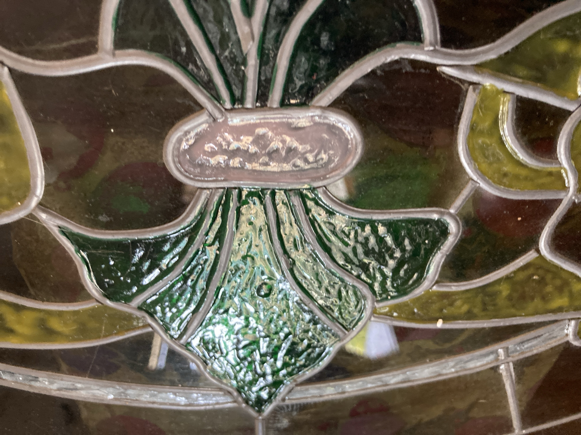 The image shows a close-up view of stained glass, featuring a green, textured leaf-like shape on the left, with a light-colored, oval inset in the center. The stained glass is bordered by thin, metallic strips. The background consists of various panes of colored glass in shades of green, yellow, and dark brown, with some translucent areas. The overall impression is of a decorative element, possibly part of a window or lamp. There are no people, discernible objects or actions visible in the image, nor is there any information about the city of Sabadell, Spain.