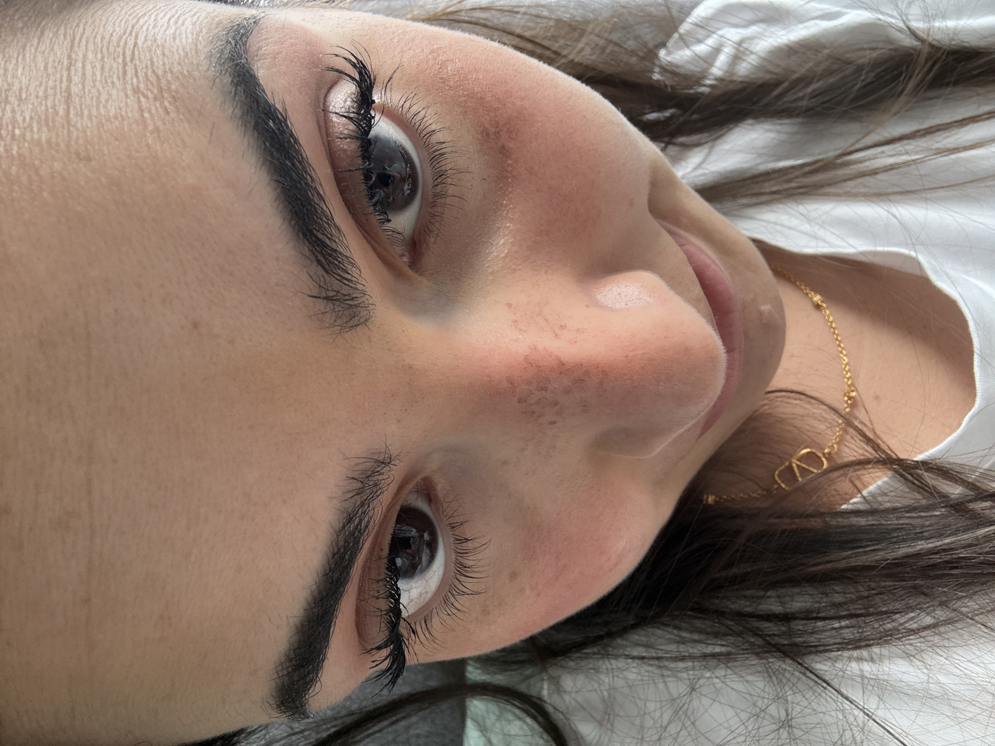 A close-up shot of a woman's face. She has dark hair and brown eyes with enhanced eyelashes. Her eyebrows are thick and well-defined. Her skin has some freckles or blemishes. She wears a delicate gold necklace with a "V" shaped pendant. She is lying down on a white surface. The location, San Bernardino Tlaxcalancingo, Mexico, is not discernable from the provided visual information.