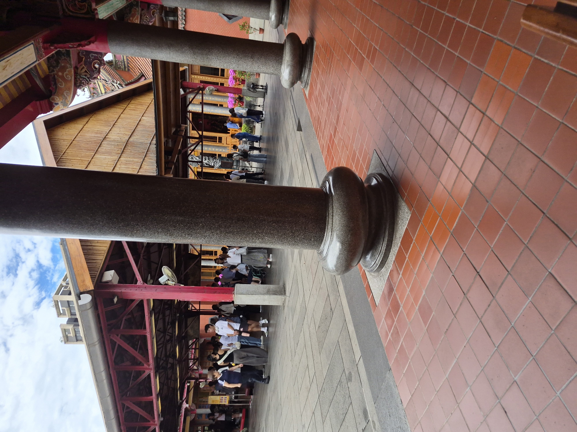 The image shows a wide-angle view of an outdoor covered walkway with a tiled floor. Large, round, stone pillars with decorative bases are prominent in the foreground and midground. Beyond the pillars, a group of people are standing and walking. The architecture suggests a temple or traditional building, with a red, open-beam structure supporting a wooden roof. The background includes a glimpse of a building with a bamboo-like wall and another structure with traditional Asian roof details. The floor is paved with reddish-brown square tiles. The sky above is partly cloudy. The scene appears to be within the Longshan Temple complex in Taipei, Taiwan, a popular tourist destination and active place of worship.