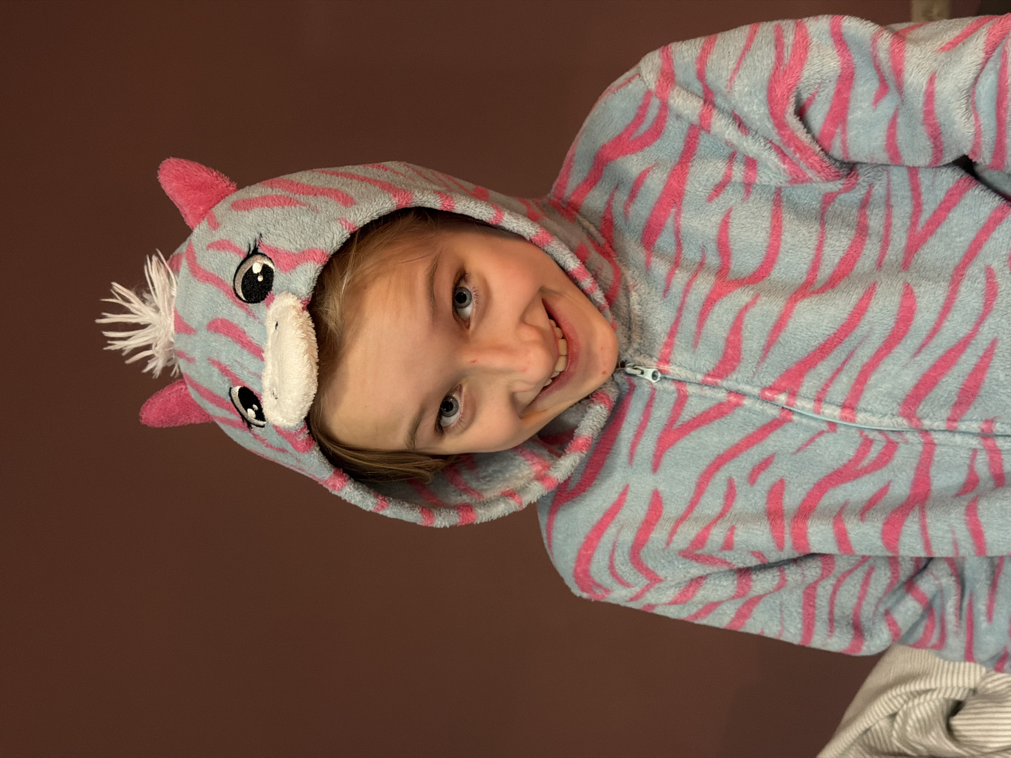 A child, around 8-10 years old, is wearing a blue and pink zebra-striped hooded onesie pajamas. The hood has ears and a unicorn horn on it. The child is smiling and tilted to the left. The background is a solid brown color, and there is a small portion of white striped fabric visible at the bottom right. The image was taken indoors.
There is no information to conclude this image was taken in Dreiliņi, Latvia.